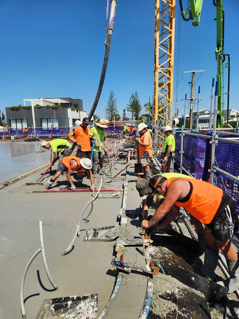 Construction workers laying concrete on a building site with machinery, safety gear, and blue fencing under clear blue sky.