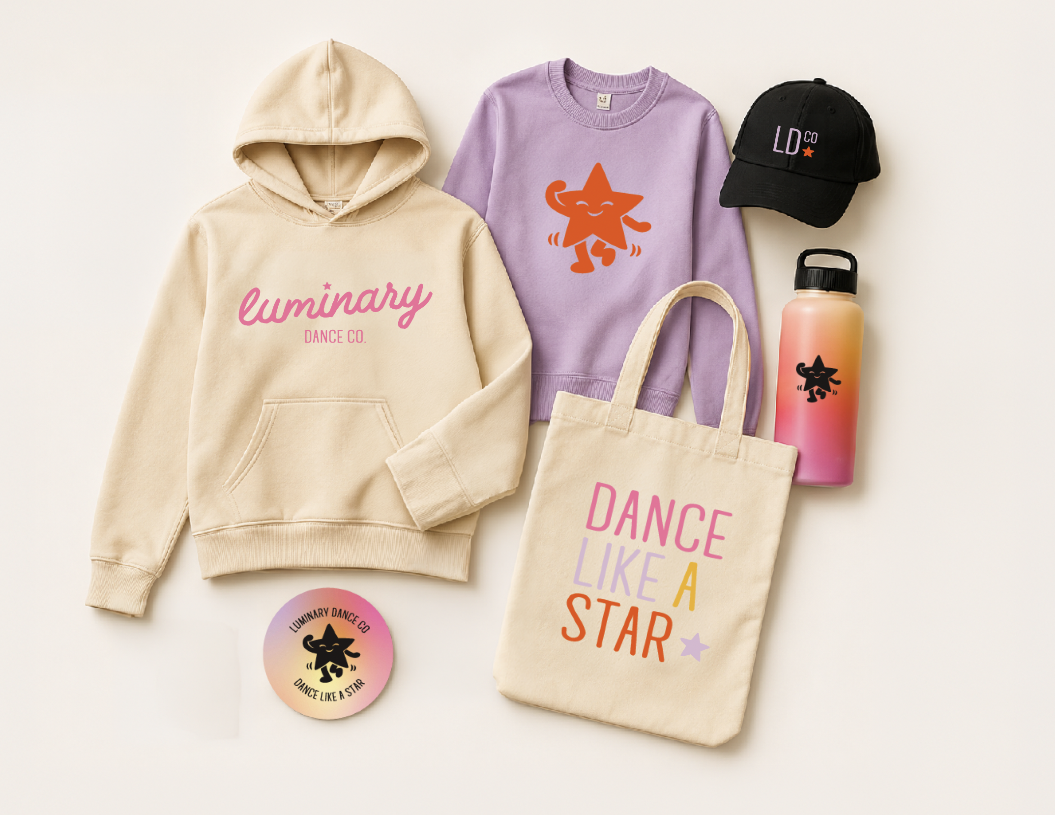 Popular clothing and accessories with colorful text and designs, including a beige hoodie with pink 'Luminary Dance Co.', a lavender sweatshirt with an orange dancing star character, a black cap with 'LD CO' and a star, a pink and orange water bottle with a black star logo, and a beige tote bag, all arranged on a white background.