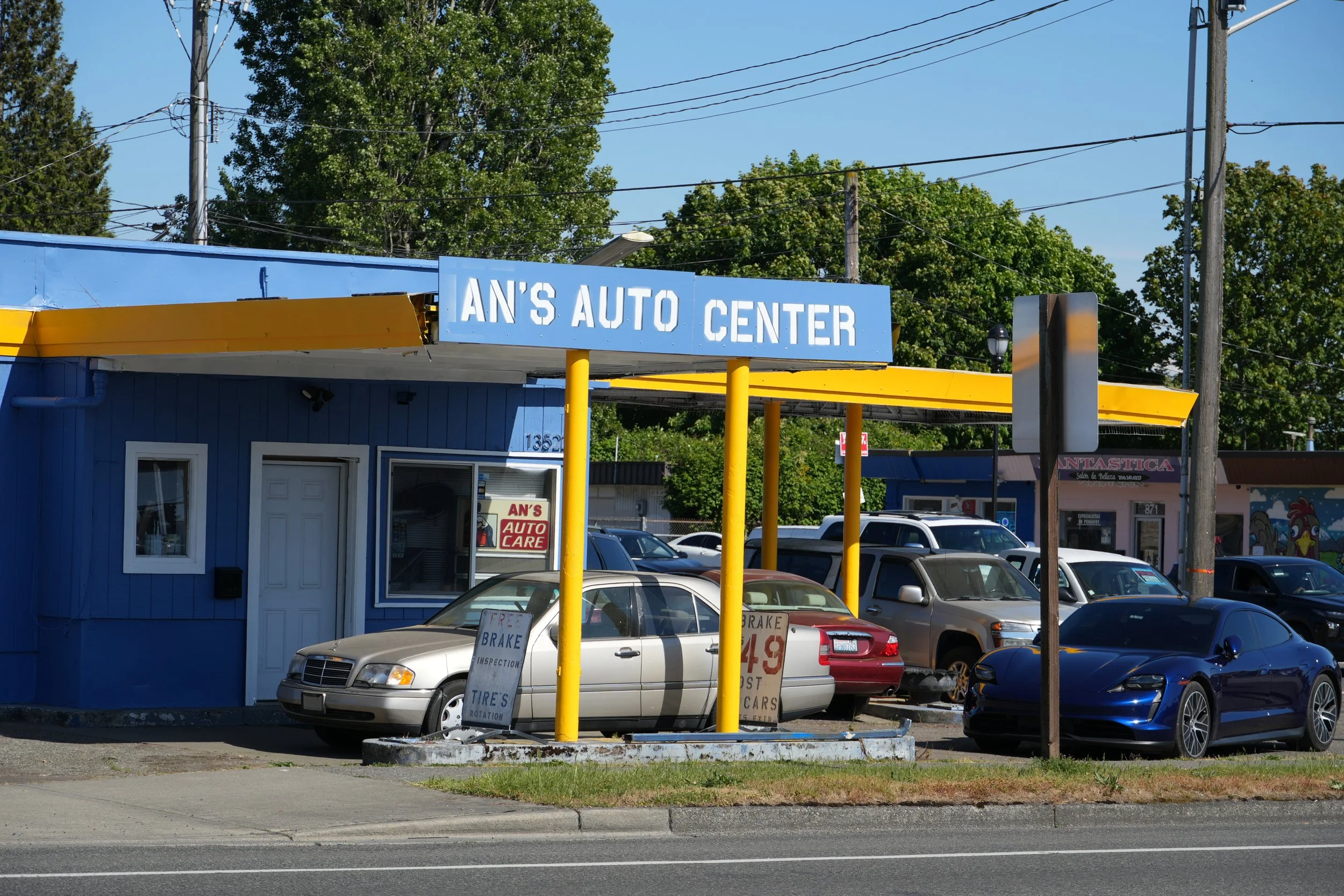 Small blue and yellow auto repair shop with cars parked outside. The storefront has signs advertising free inspections, tire rotations, and checkups, with some signs visible through the windows.