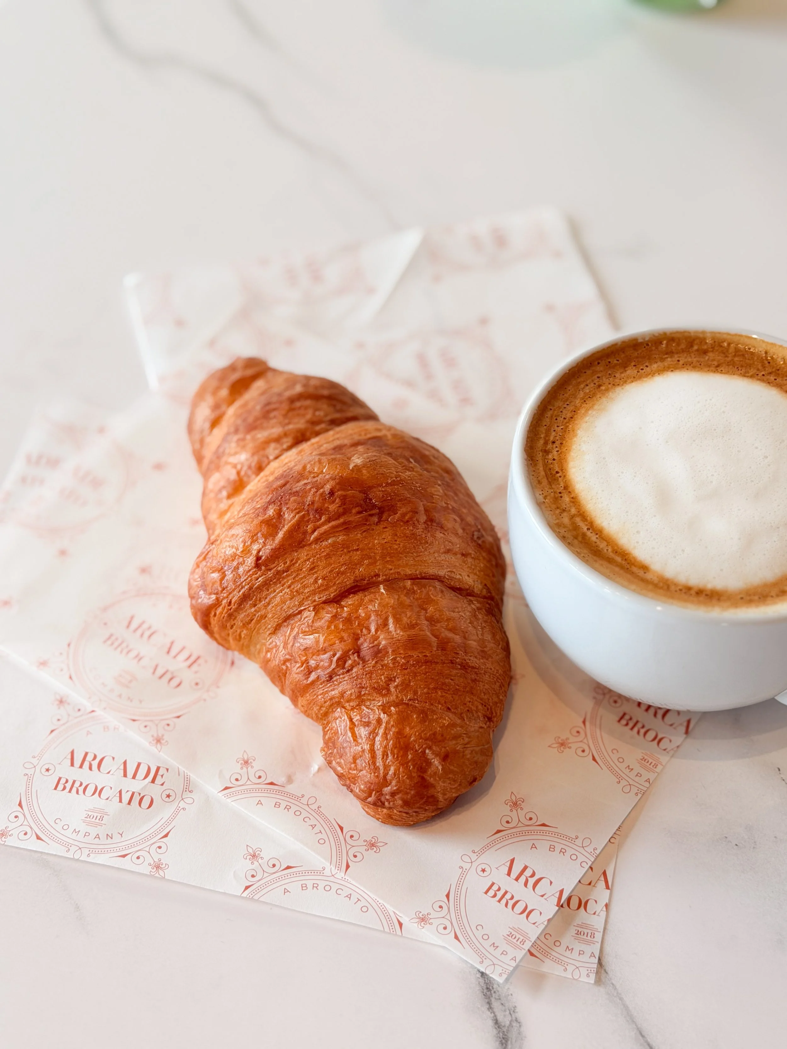 A croissant and a cup of coffee on a white marble surface with branded paper underneath.