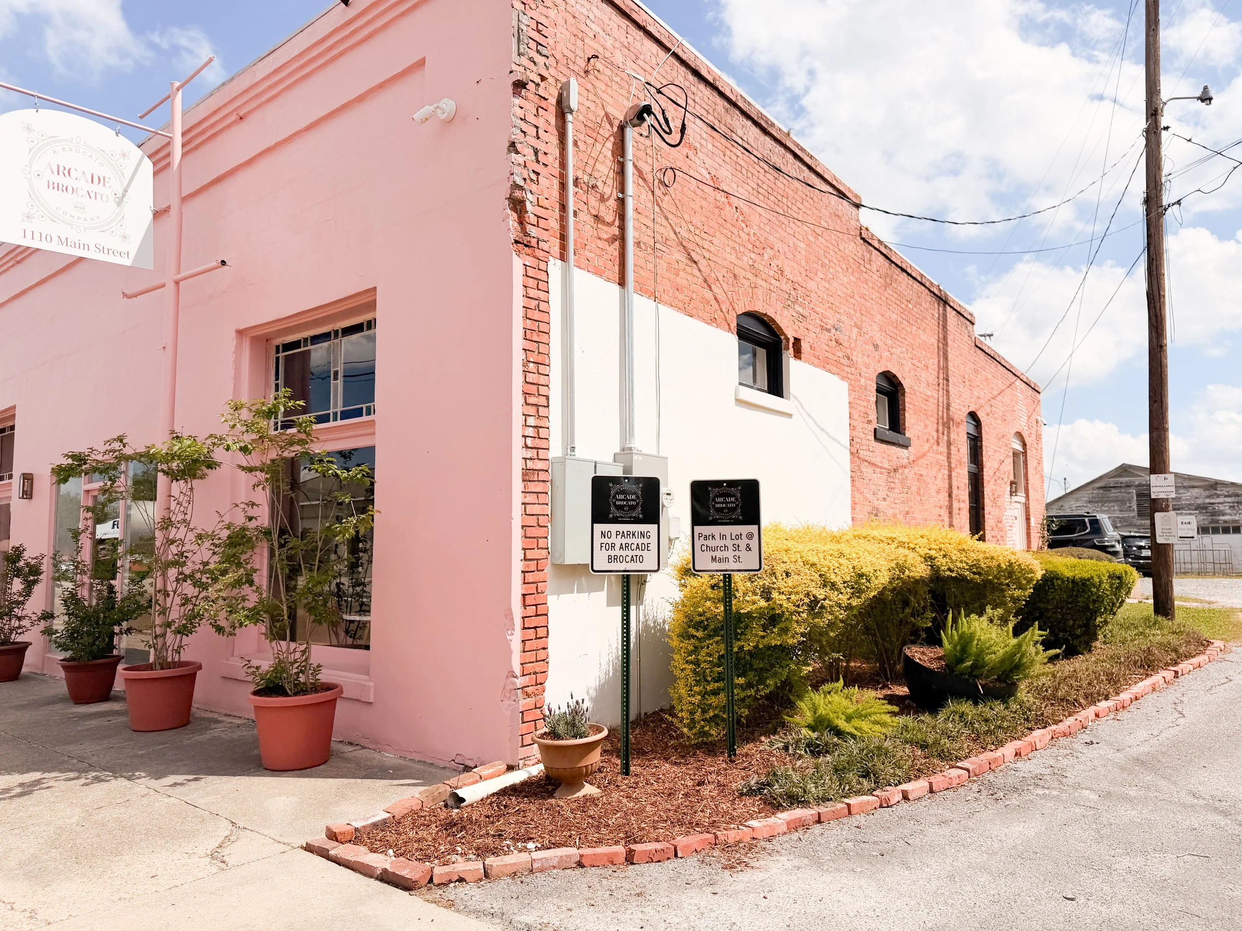 Corner of a pink and red brick building with plants in pots and bushes outside. There are two parking signs in front of the building with parking info
