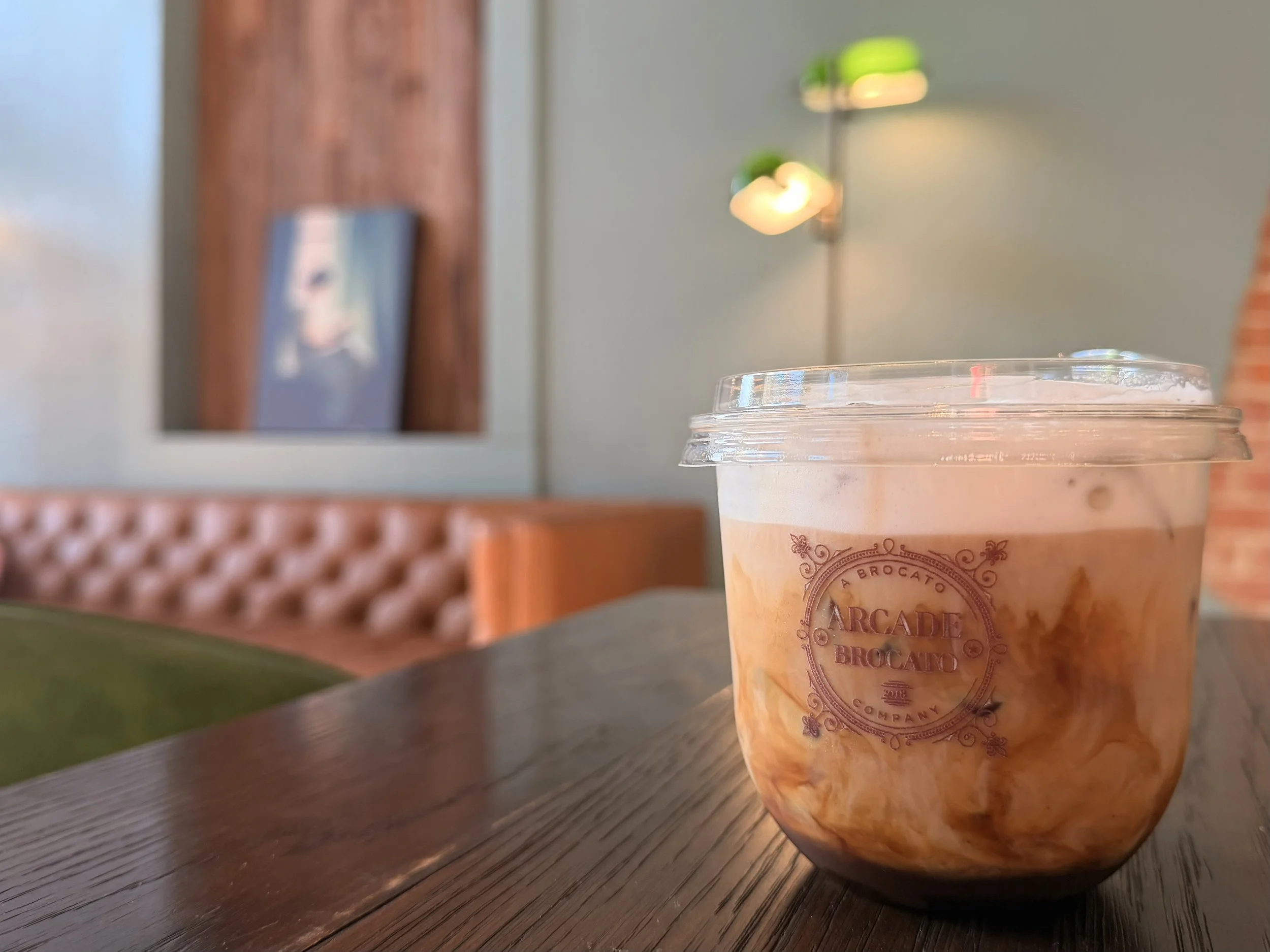 A plastic cup with a clear lid containing a layered iced coffee drink with milk and coffee, placed on a wooden table inside a cafe with blurred background.