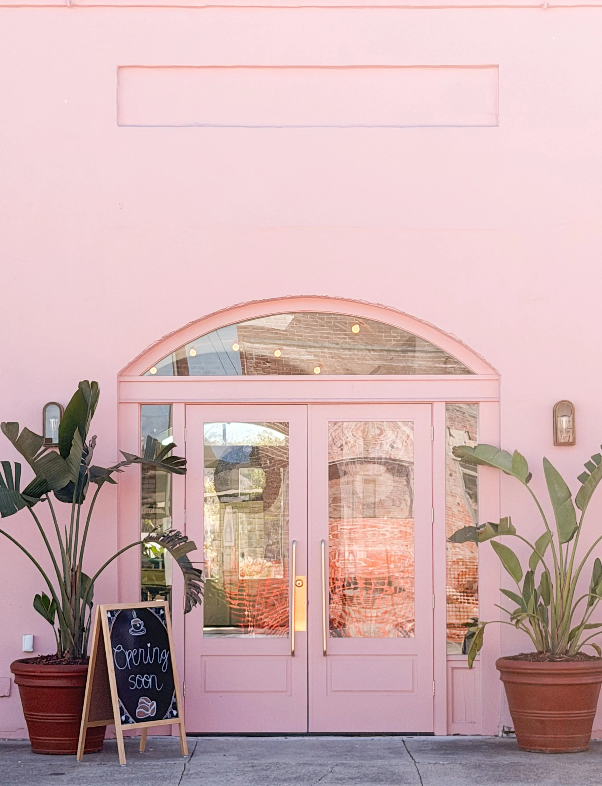 Pink storefront with glass double doors, potted plants on either side, and a chalkboard sign that says 'Opening soon' with a coffee cup drawing.