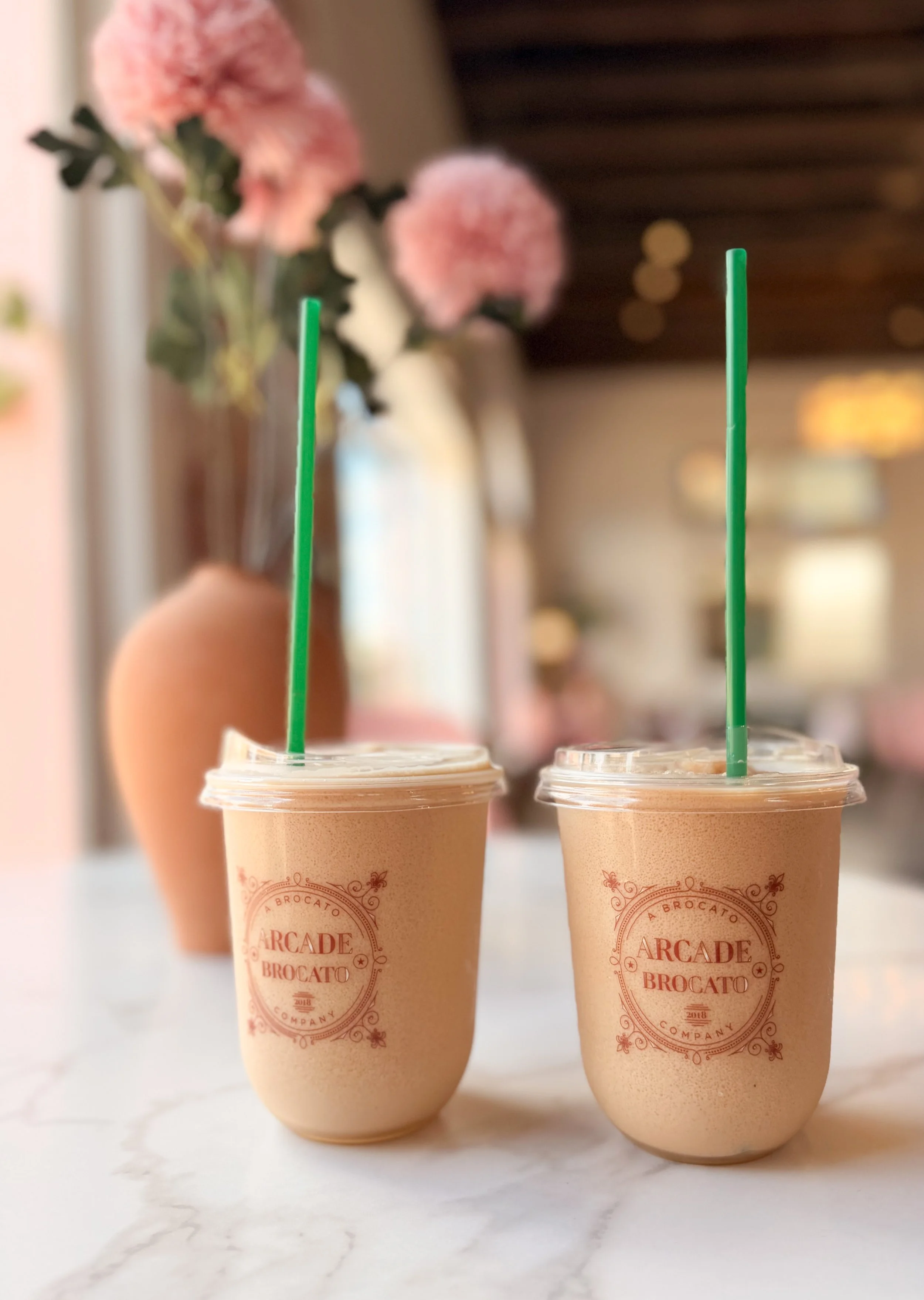 Two cups of the Arcade Freeze with green straws on a white marble table, with a pink flower arrangement in the background.