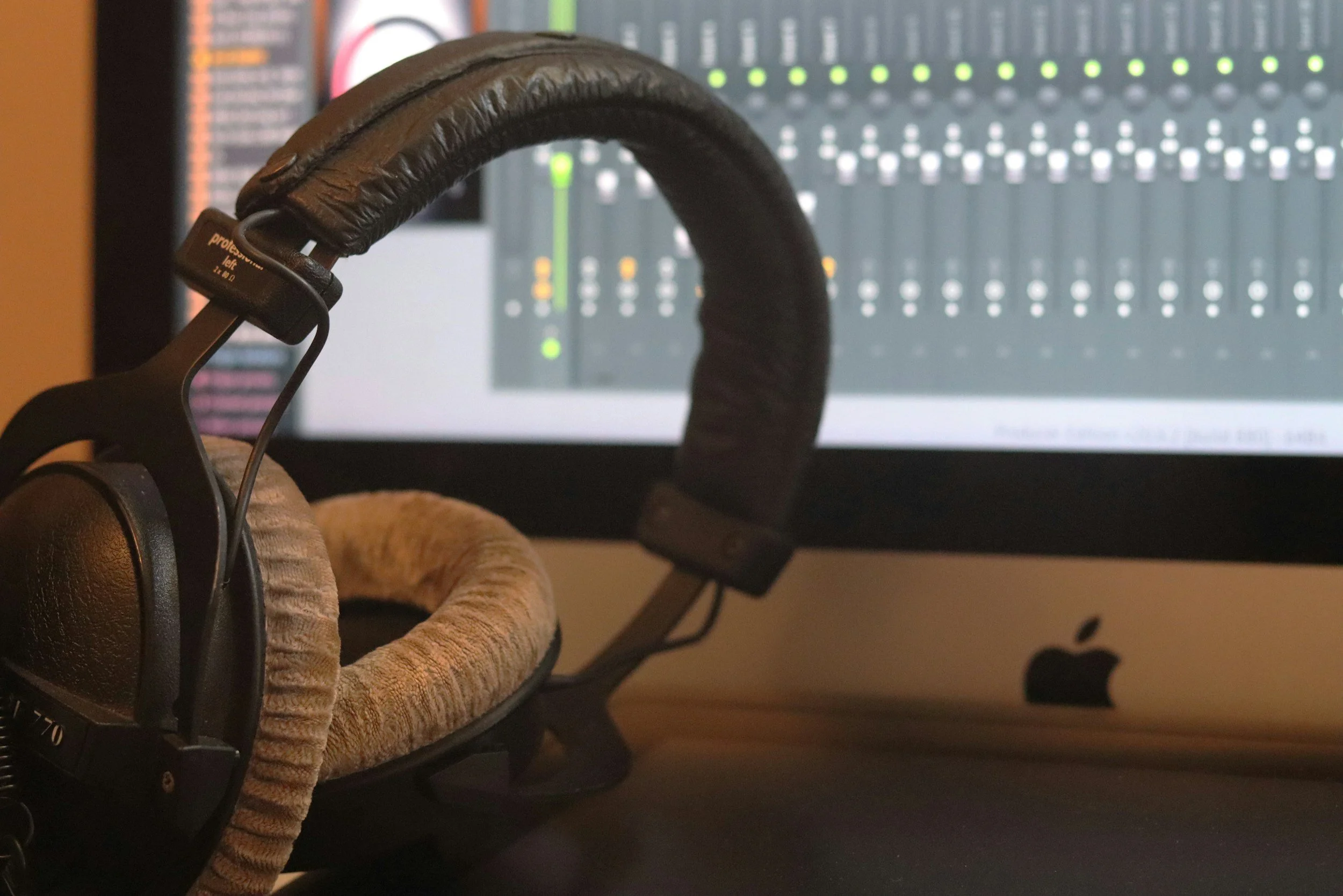 A pair of headphones resting on a beige-colored cat ear headband in front of a computer monitor with digital audio mixing software.