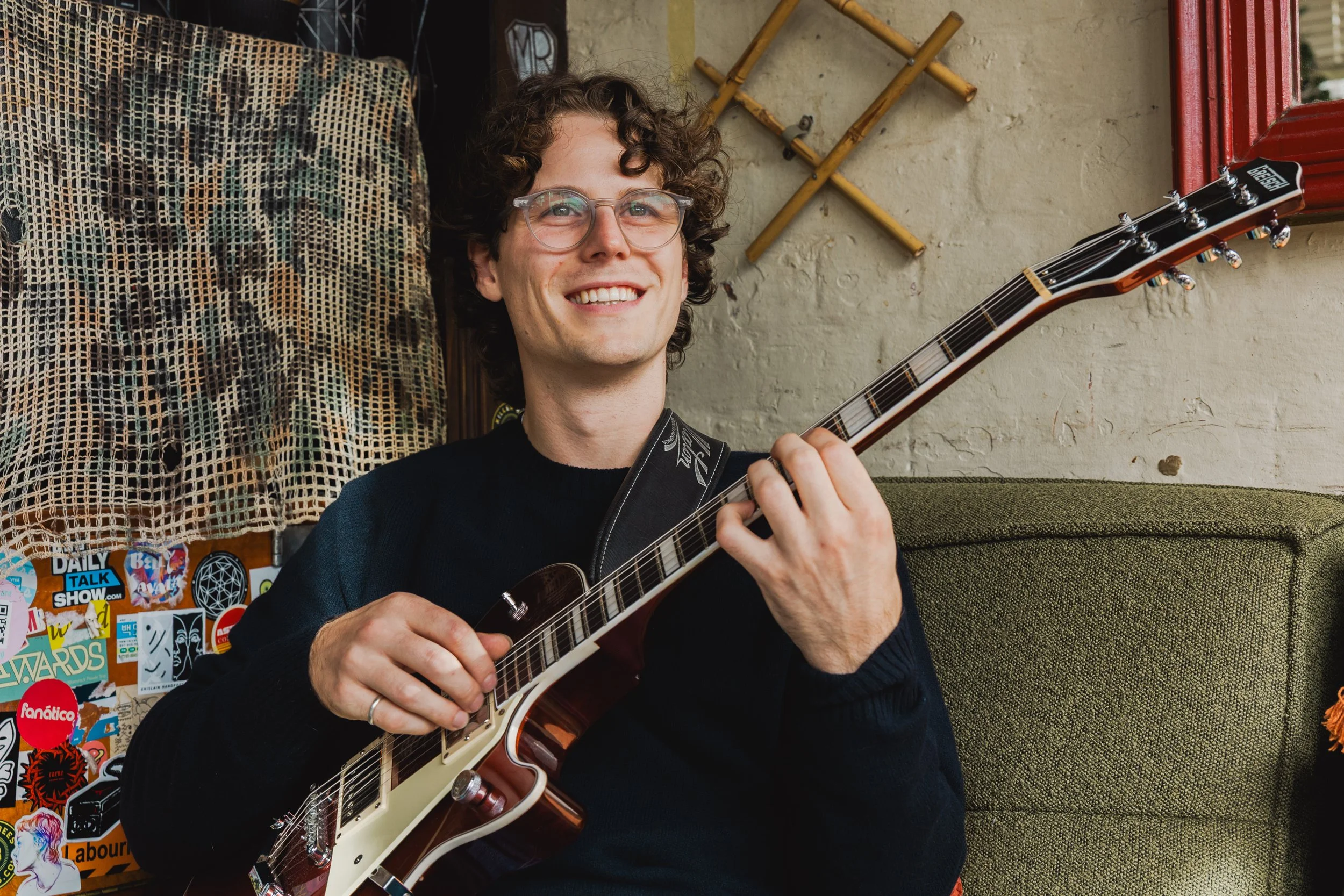 A young man from Melboure with curly hair and glasses smiling while playing an electric guitar, sitting on a green couch with a wall behind him decorated with stickers and a plant. An abstract wall hanging is visible on his left.