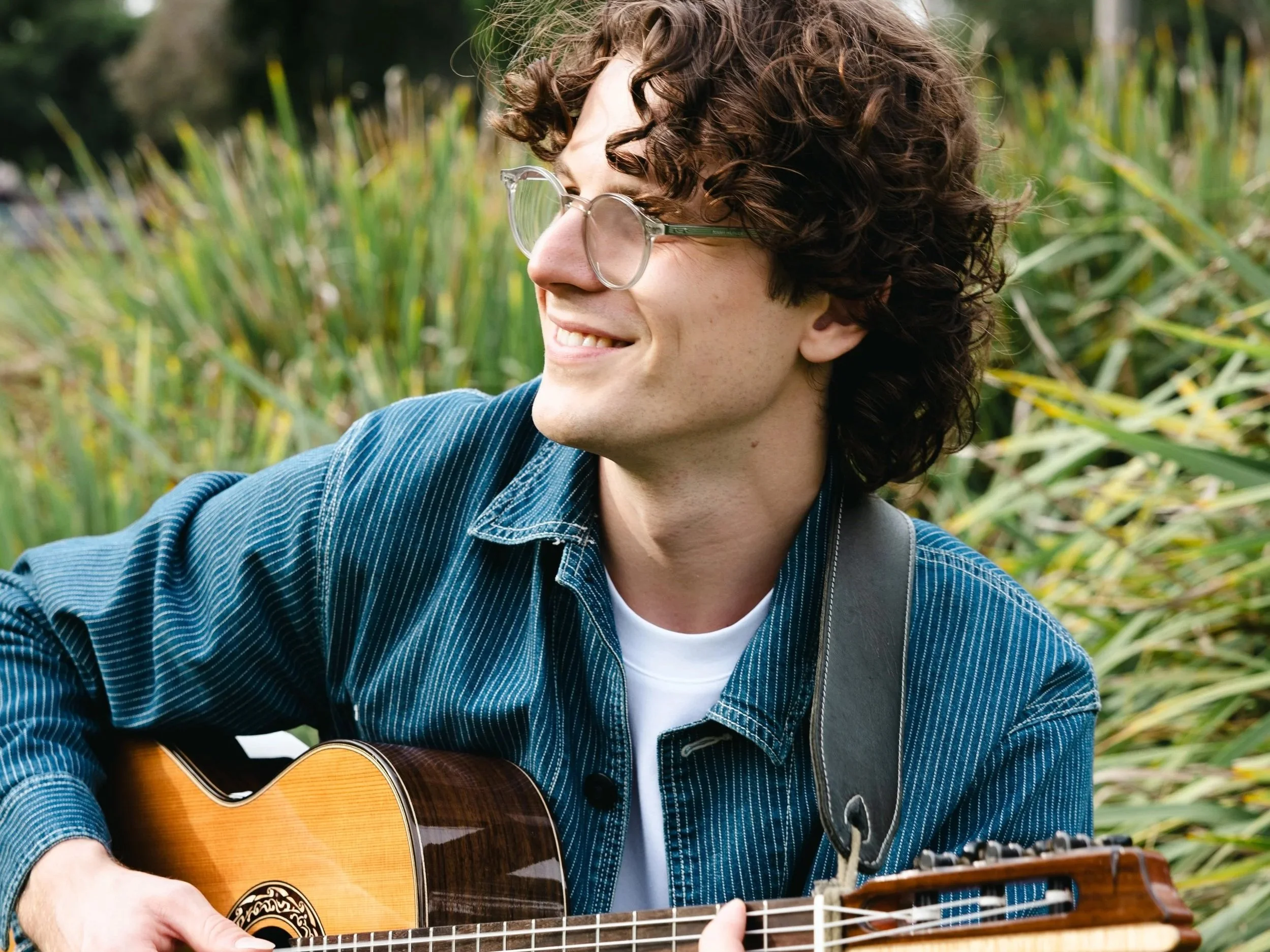 A young man from Melbourne with curly hair and glasses playing an acoustic guitar outdoors, smiling, with a backdrop of green plants.