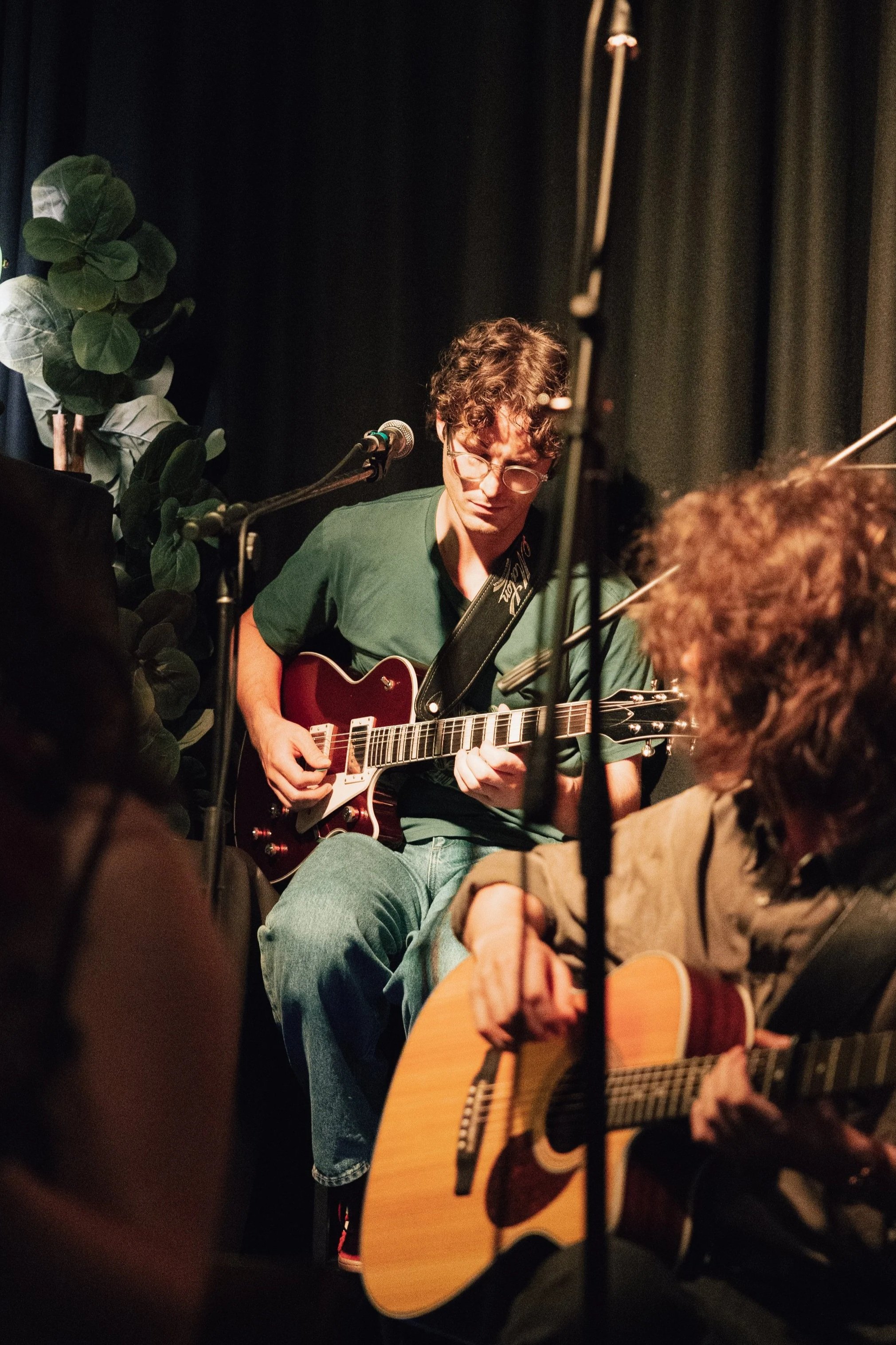 A young person in Melbourne with curly hair and glasses playing an electric guitar on stage, surrounded by microphones and audience members, with a dark curtain backdrop and some green foliage on the side.