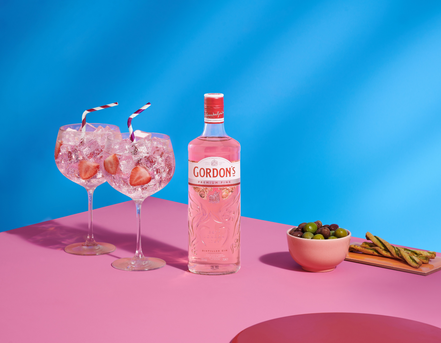 A pink bottle of Gordon's Premium Pink gin, two pink cocktails with strawberries and striped straws, a bowl of green and purple olives, and a plate of breadsticks on a pink surface with a blue background.