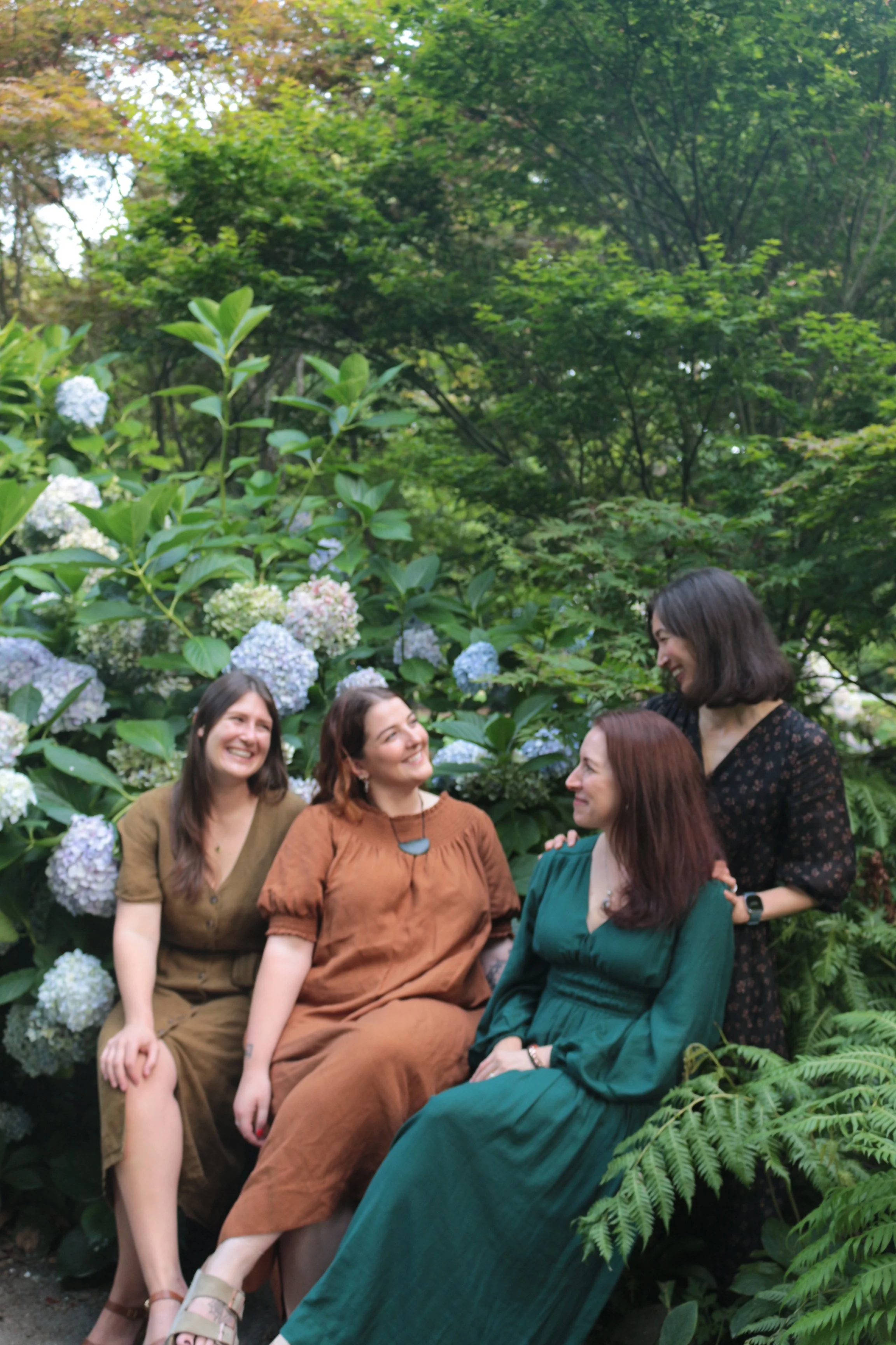 Four women sitting and standing near hydrangea bushes in a lush, green, outdoor garden, smiling and interacting with each other.
