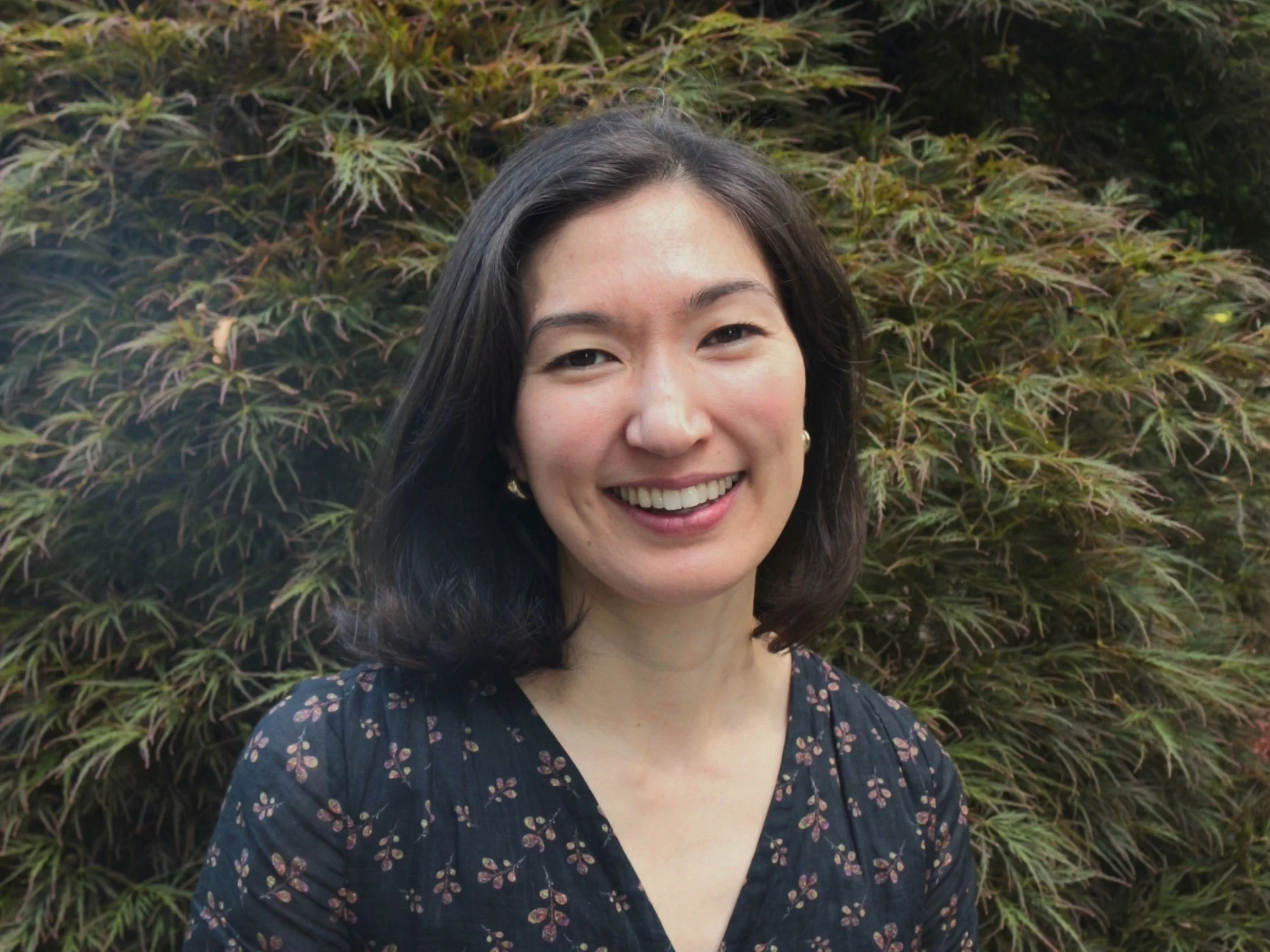 A smiling woman with short dark hair, wearing a dark floral dress, standing in front of leafy bushes.