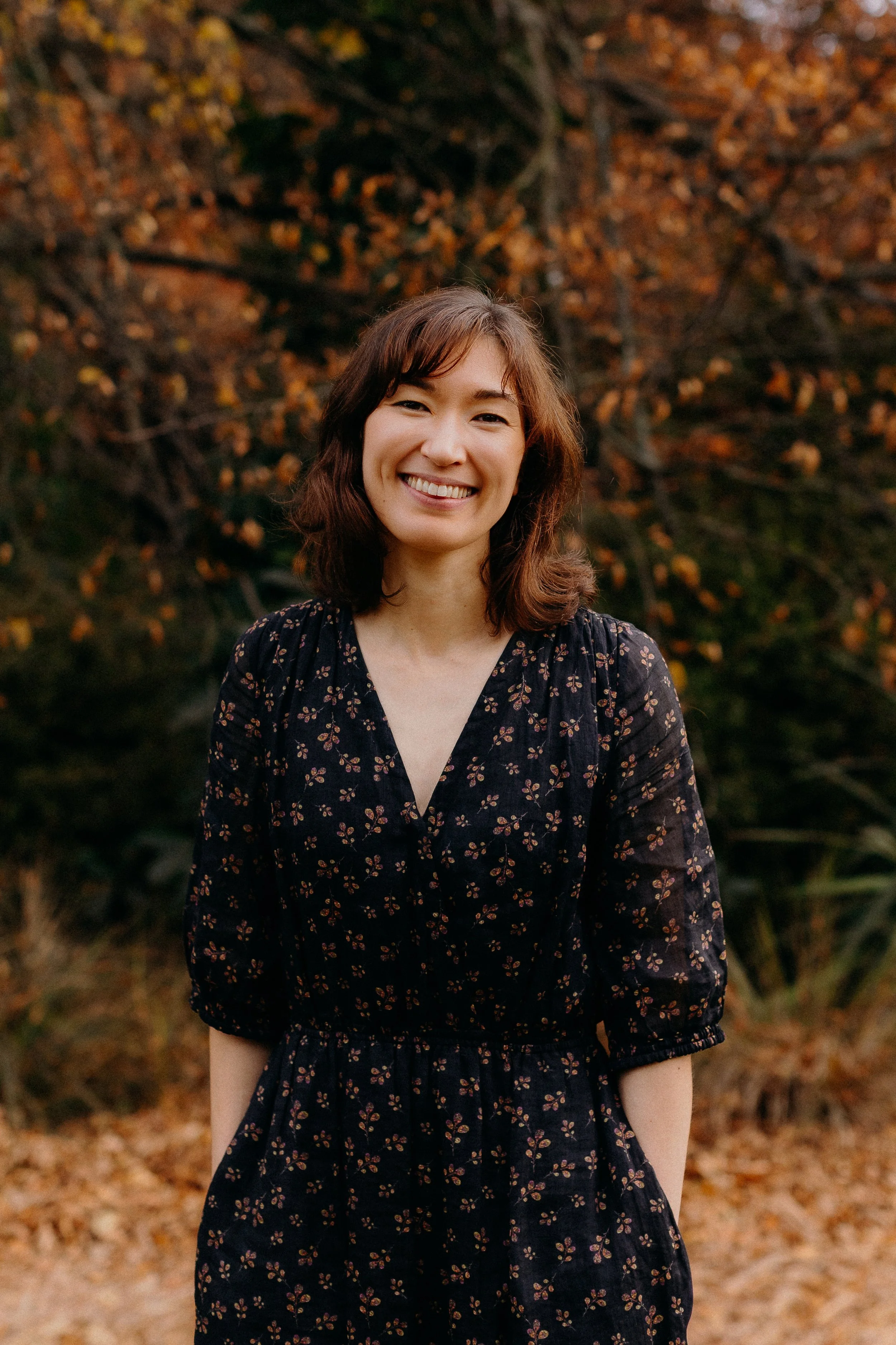 A woman with shoulder-length dark hair, smiling in front of green foliage with reddish tips.