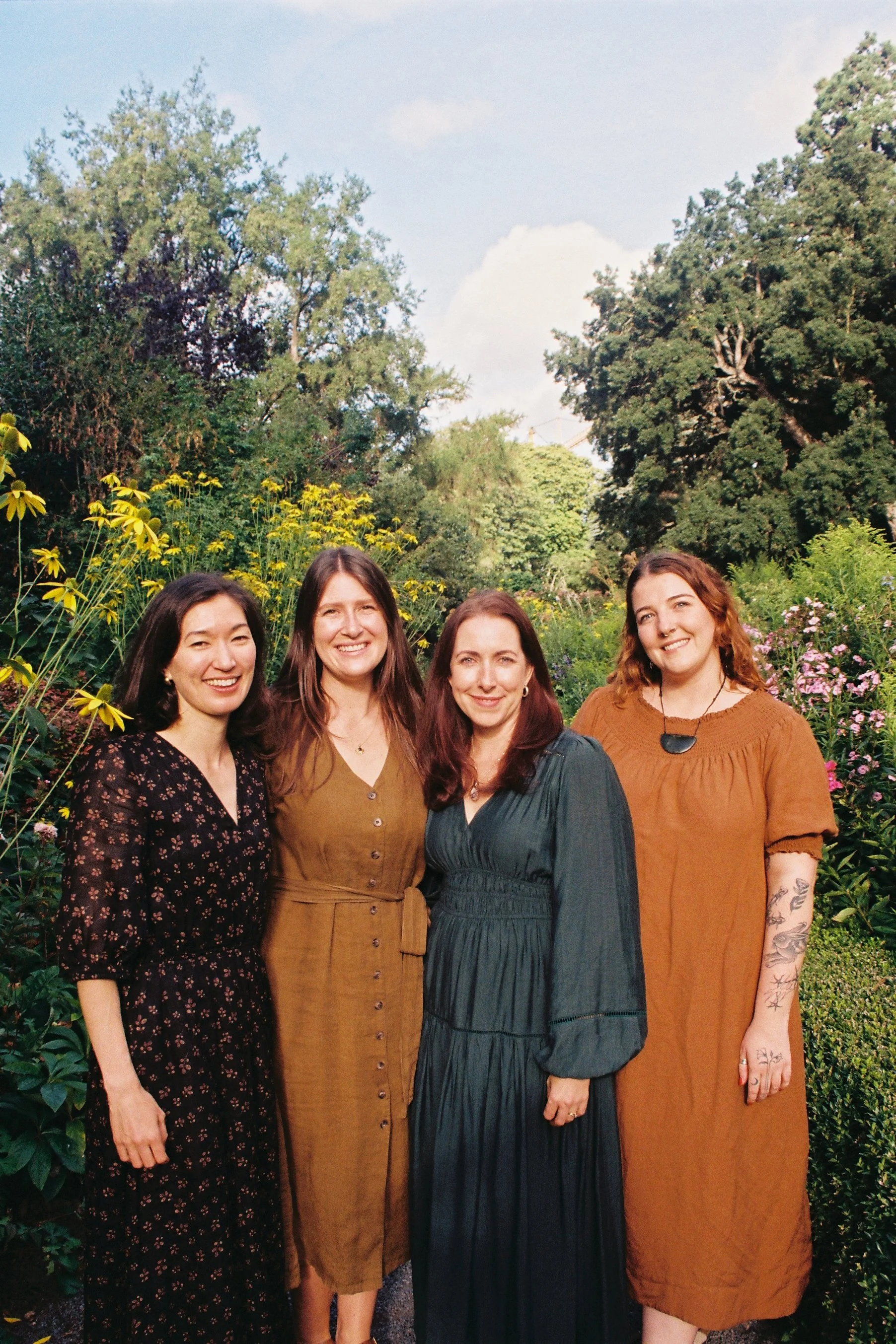 Four women standing together outdoors in a garden with trees and flowers in the background, smiling at the camera.