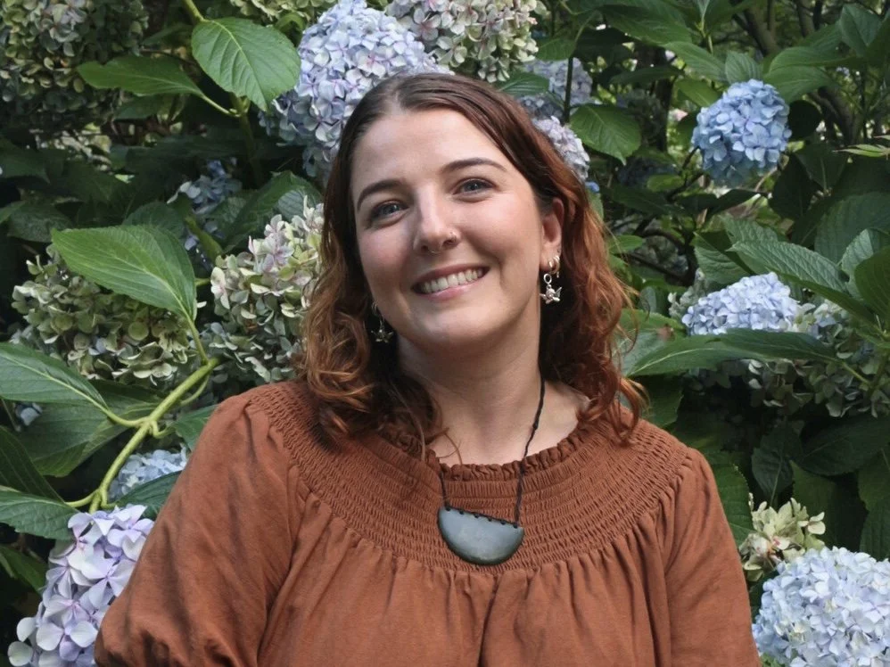 A woman with red hair smiling in front of green leaves and light purple hydrangea flowers.