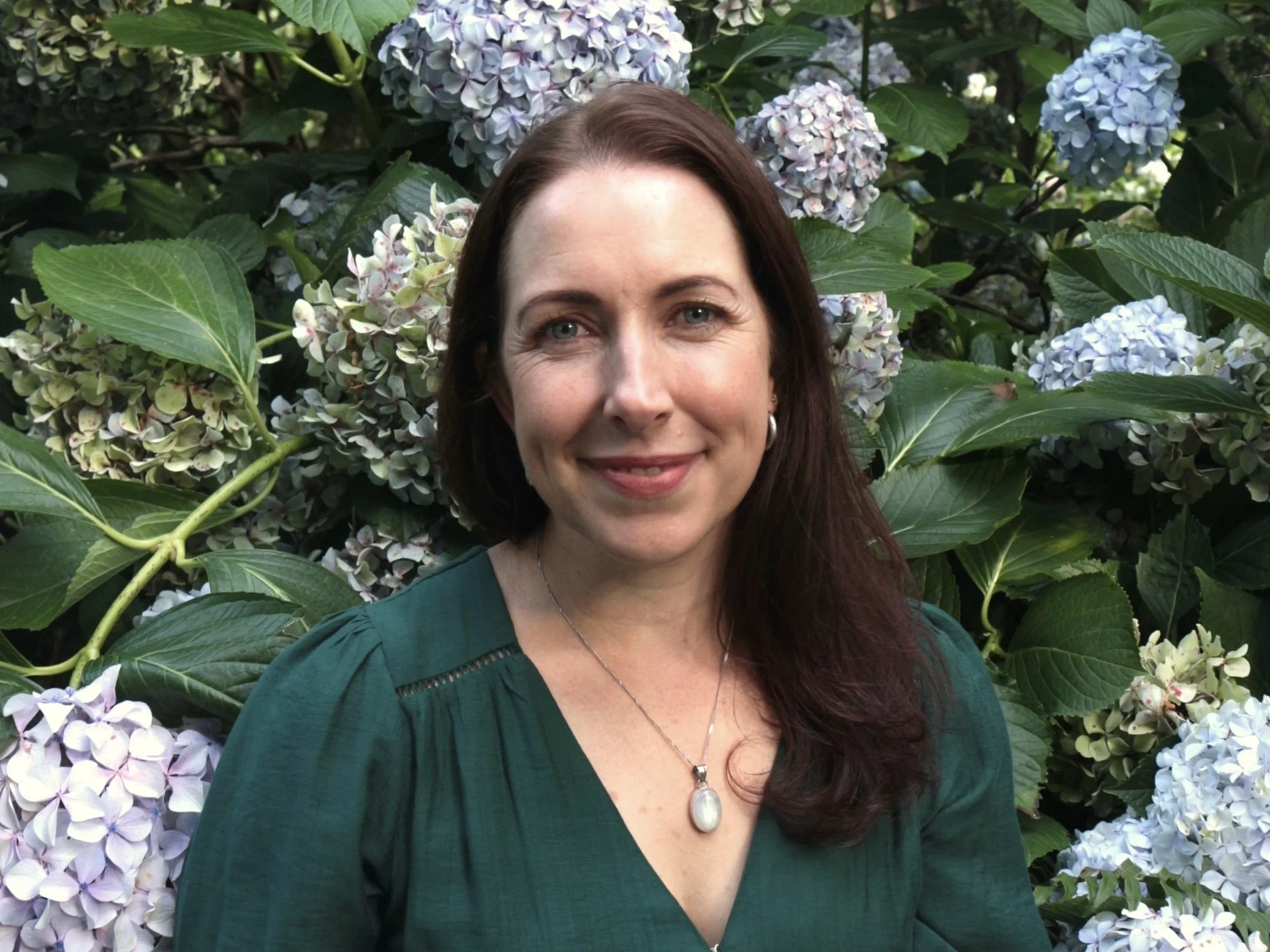 A woman with brown hair and green eyes smiling in front of hydrangea flowers in a garden.