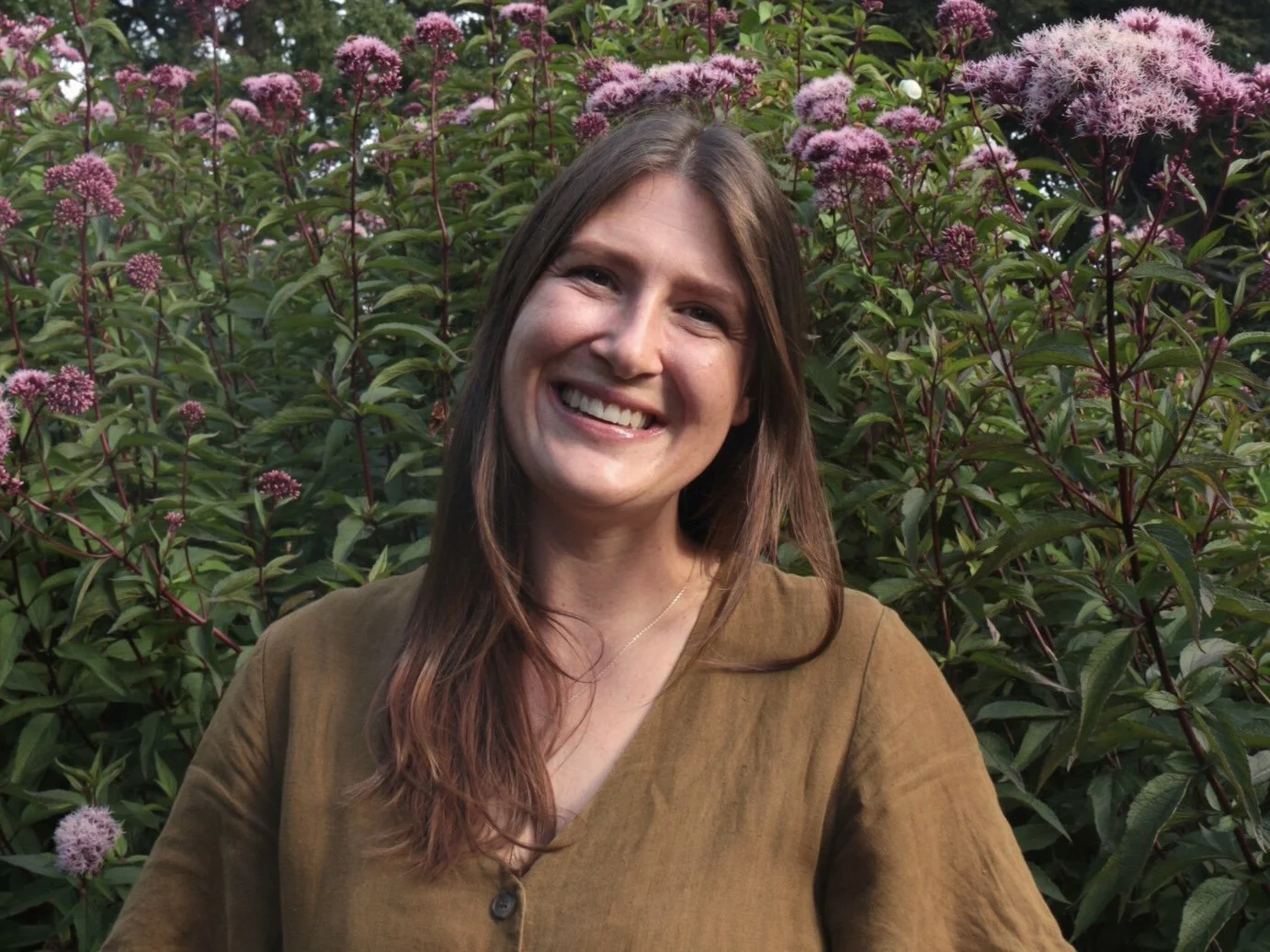 A woman smiling outdoors with pink and purple flowers in the background.