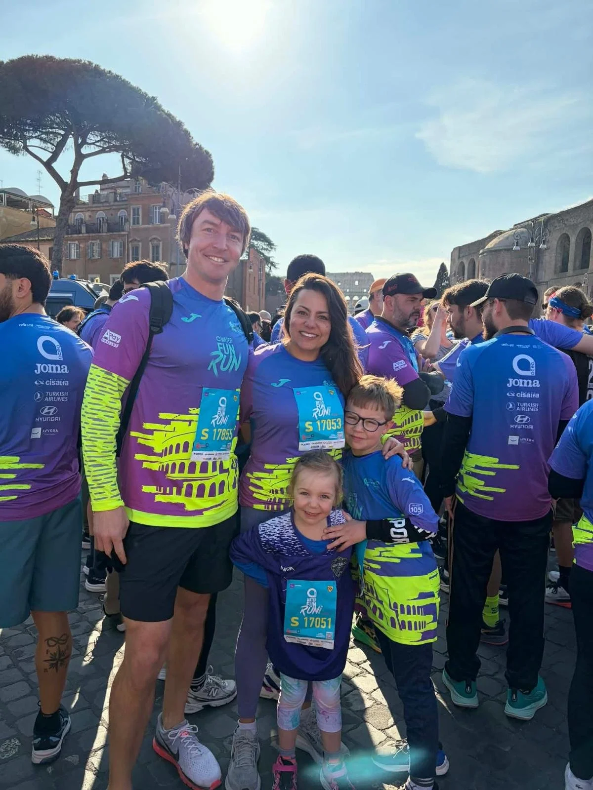 A group of runners and kids in colorful race shirts at a race event, with historic buildings and trees in the background, under a sunny sky.