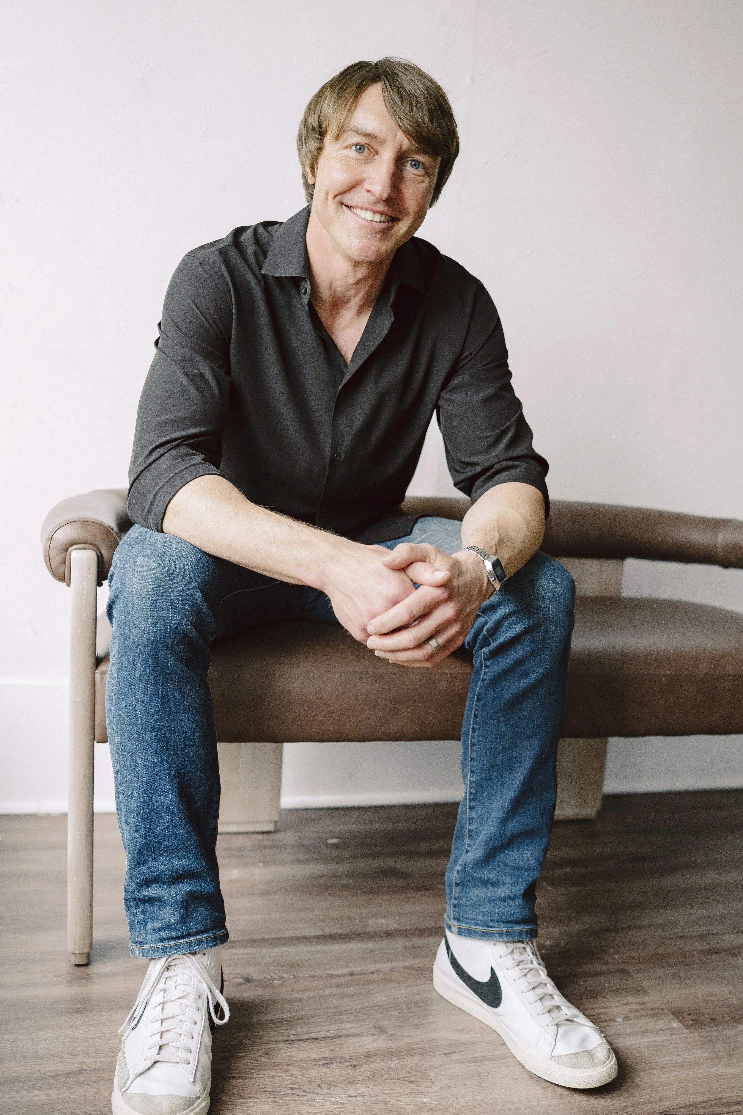 A man with light brown hair, blue eyes, and fair skin, smiling, seated on a beige wooden bench with a neutral tone wall in the background. He's dressed in a black button-up shirt, blue jeans, and white sneakers with a green Nike swoosh. His hands are clasped together, resting on his lap.
