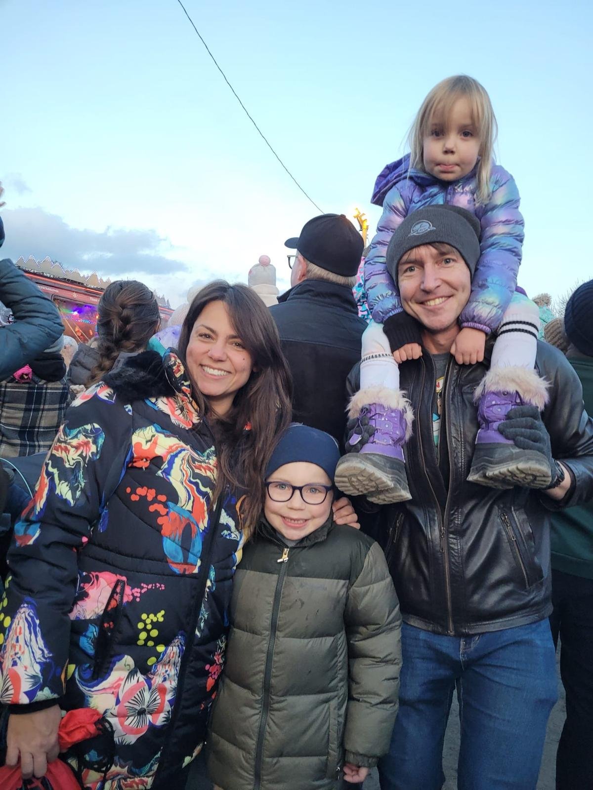A happy family of four outdoors in cold weather, with a crowd in the background and cloudy sky. The family includes a woman, a man, a young girl with glasses, and a younger girl sitting on the man's shoulders, all smiling and dressed warmly.