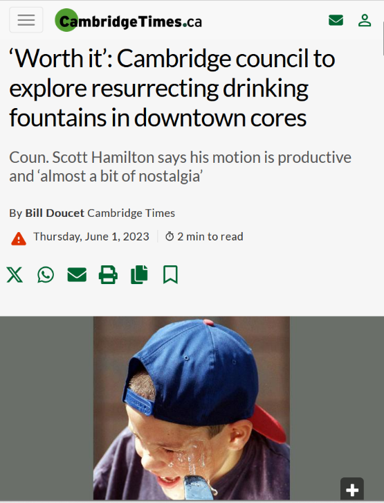 Child wearing a blue baseball cap with a red brim, drinking from a water fountain outdoors.