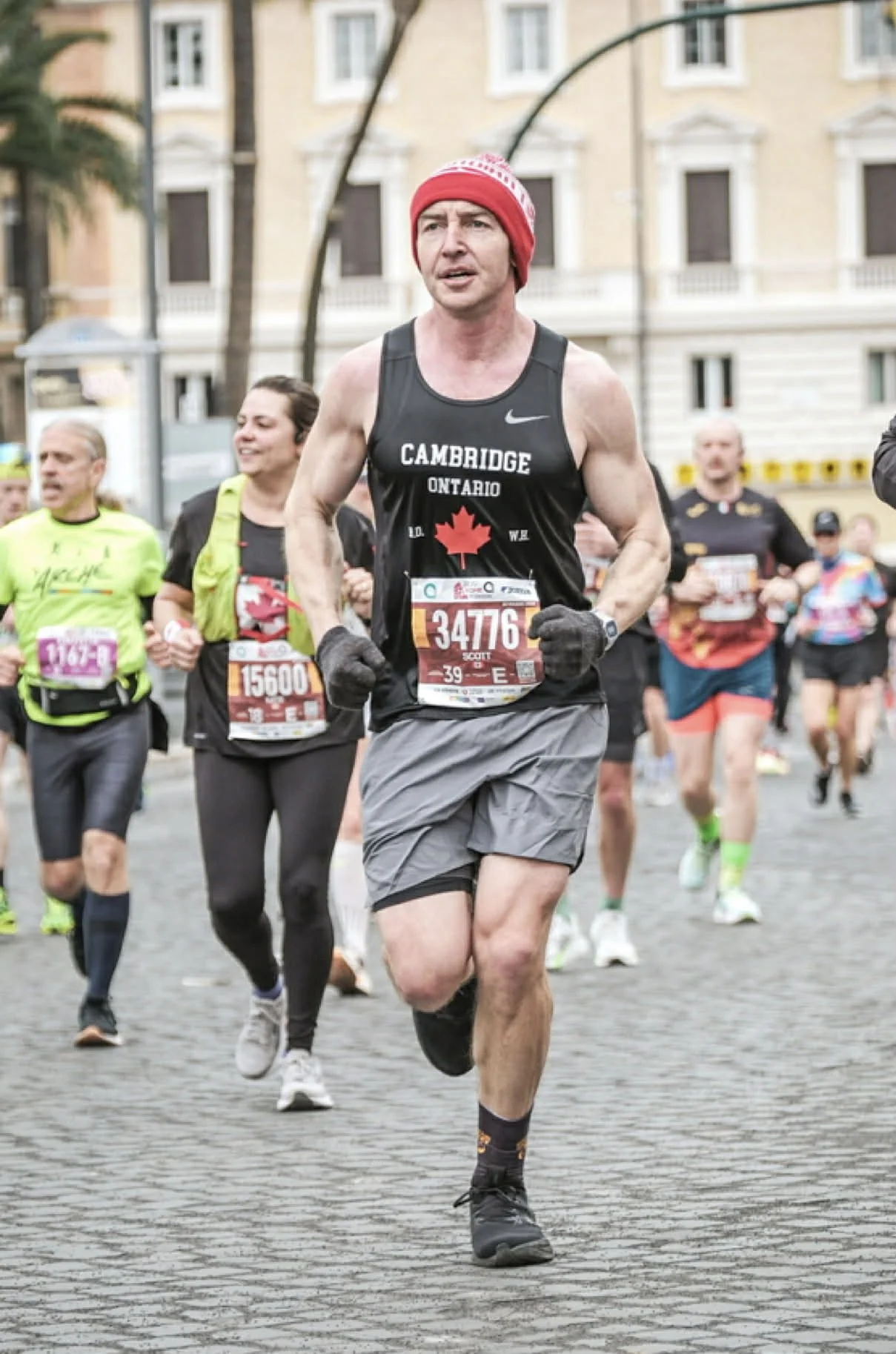 A man in a black tank top with a red maple leaf and the words 'Cambridge Ontario' runs in a marathon, surrounded by other runners on a city street.