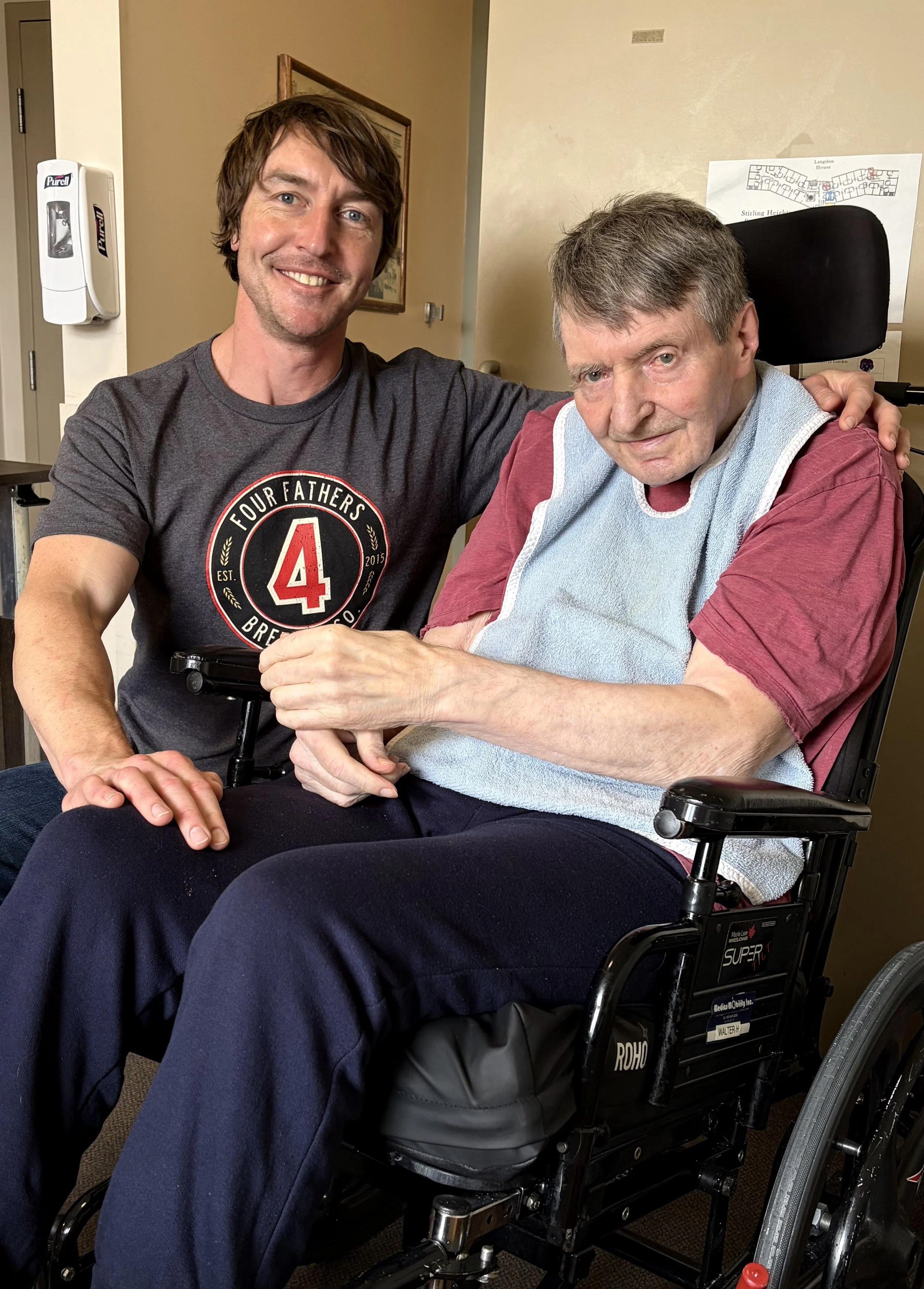 A young man with dark brown hair and a beard is smiling and sitting with an older man in a wheelchair. The older man has gray hair and is wearing a maroon shirt with a blue hospital gown. The young man's arm is around the older man's shoulder, and they are holding hands. The background shows a beige wall with a framed picture and a white hand sanitizer dispenser.