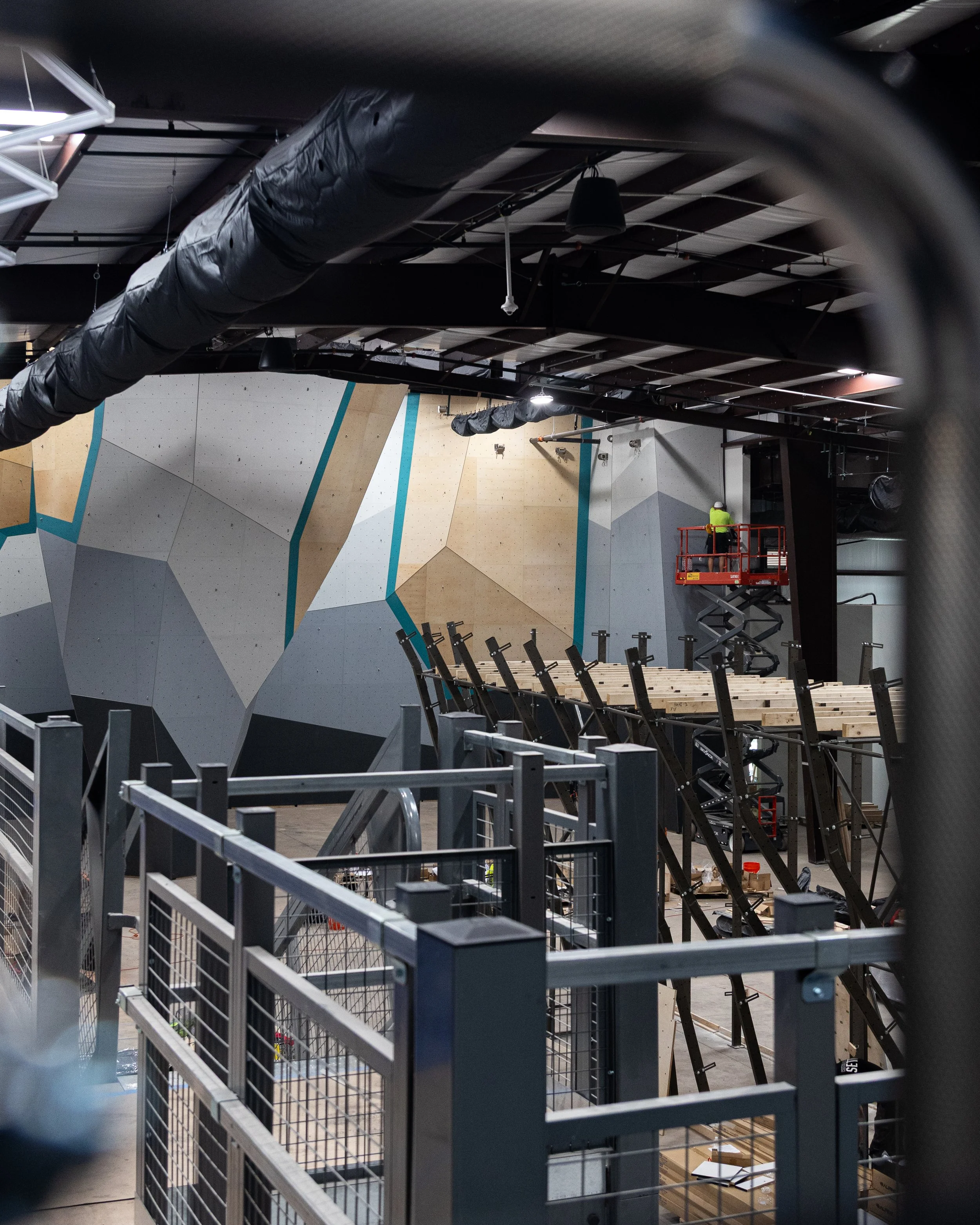 Construction workers building an indoor climbing gym with large geometric climbing walls, scaffolding, and safety barriers.
