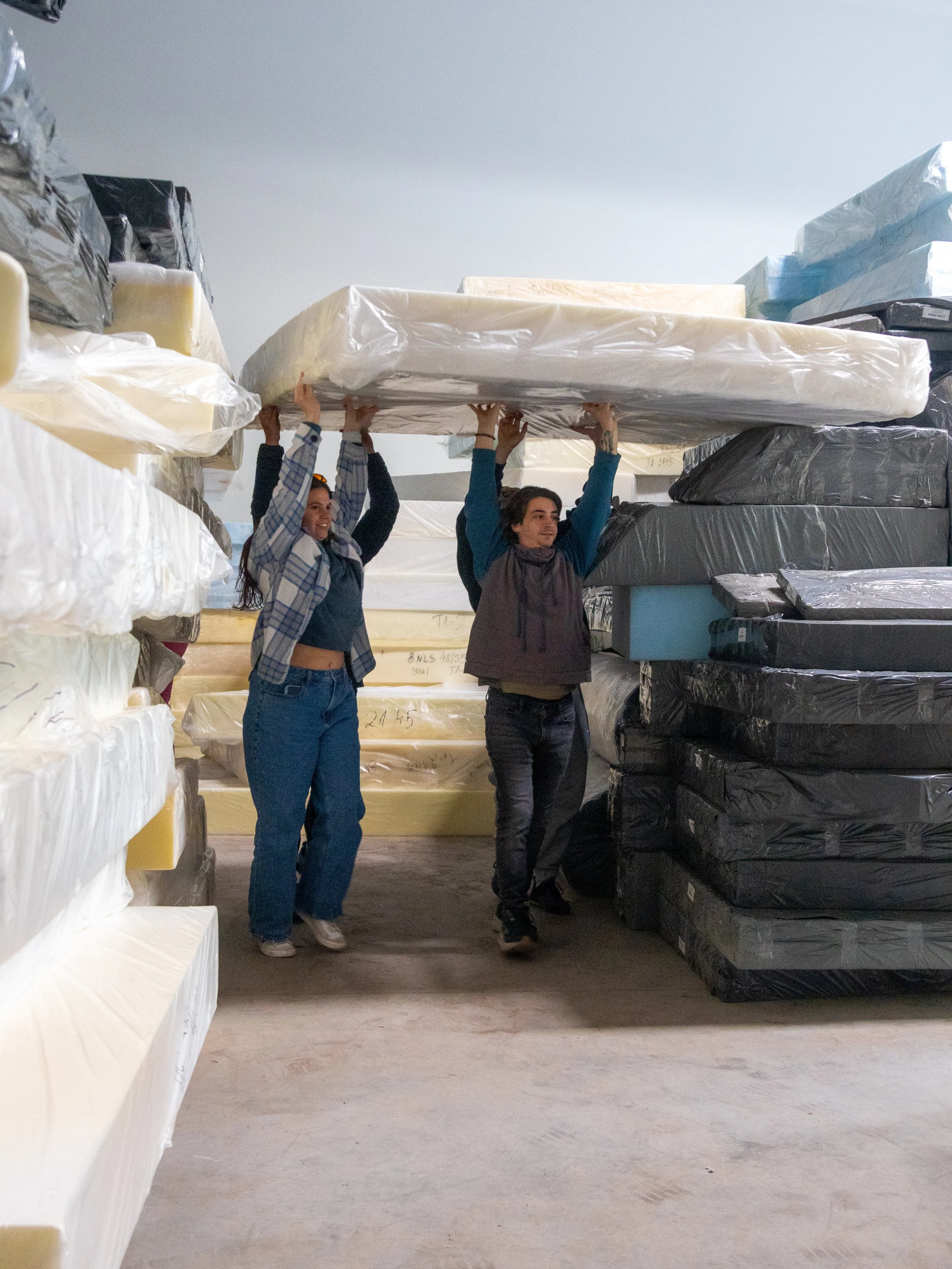 Two people carrying a mattress in a warehouse with stacked mattresses around them.