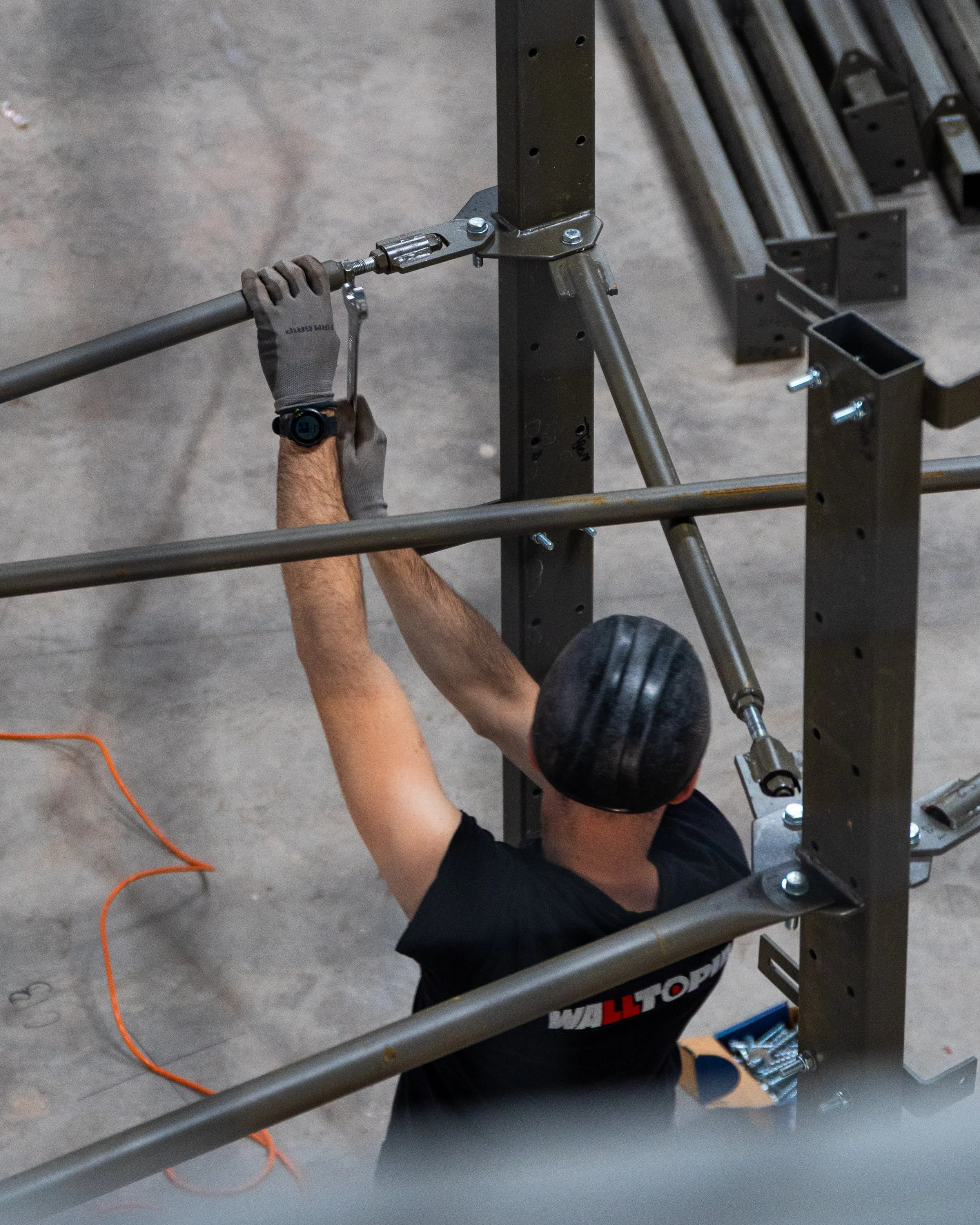 A person wearing a black shirt and a black cap is assembling steel frame parts together in a workshop or gym, with metal beams and tools around.