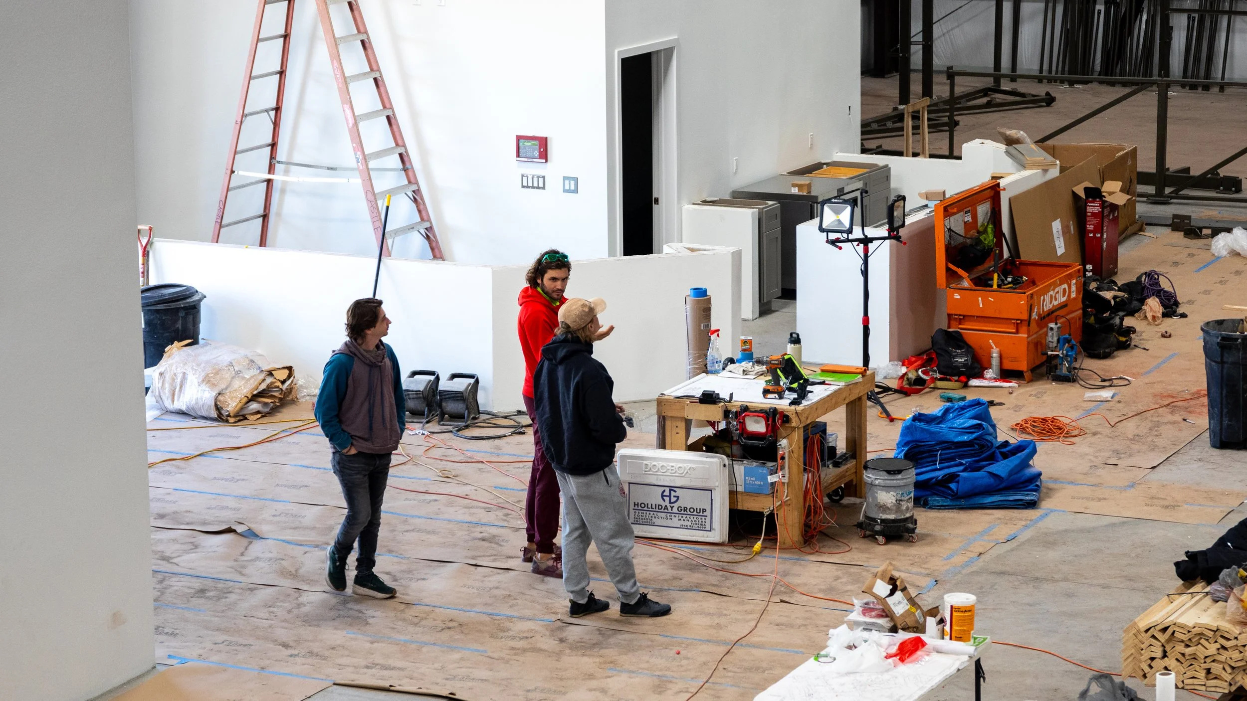Three people standing on a construction site with tools, equipment, and building materials scattered around. A large red ladder is against the white wall, and a work table with various tools is in the middle.