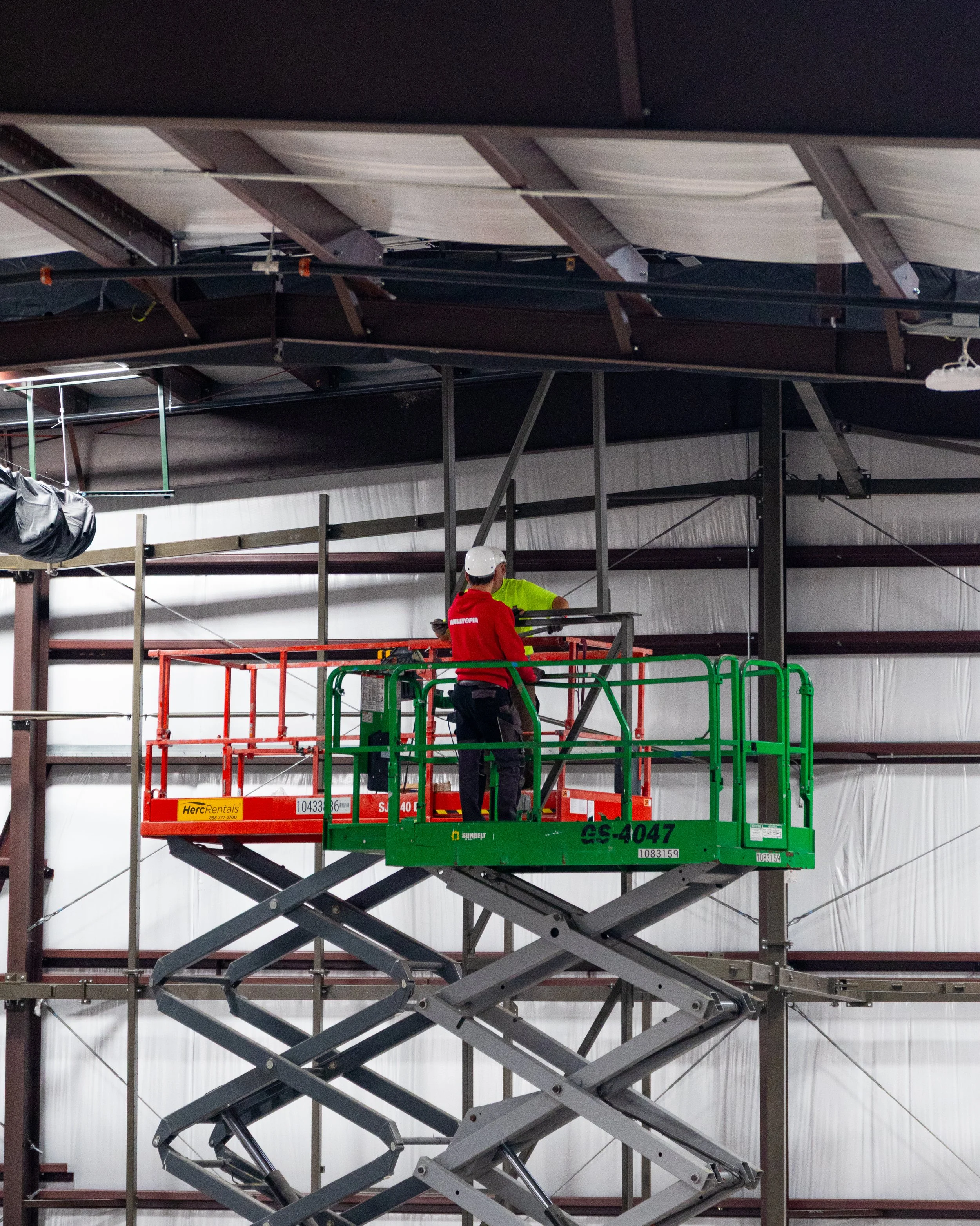 Two workers on a green and orange scissor lift inside a large metal structure under construction.