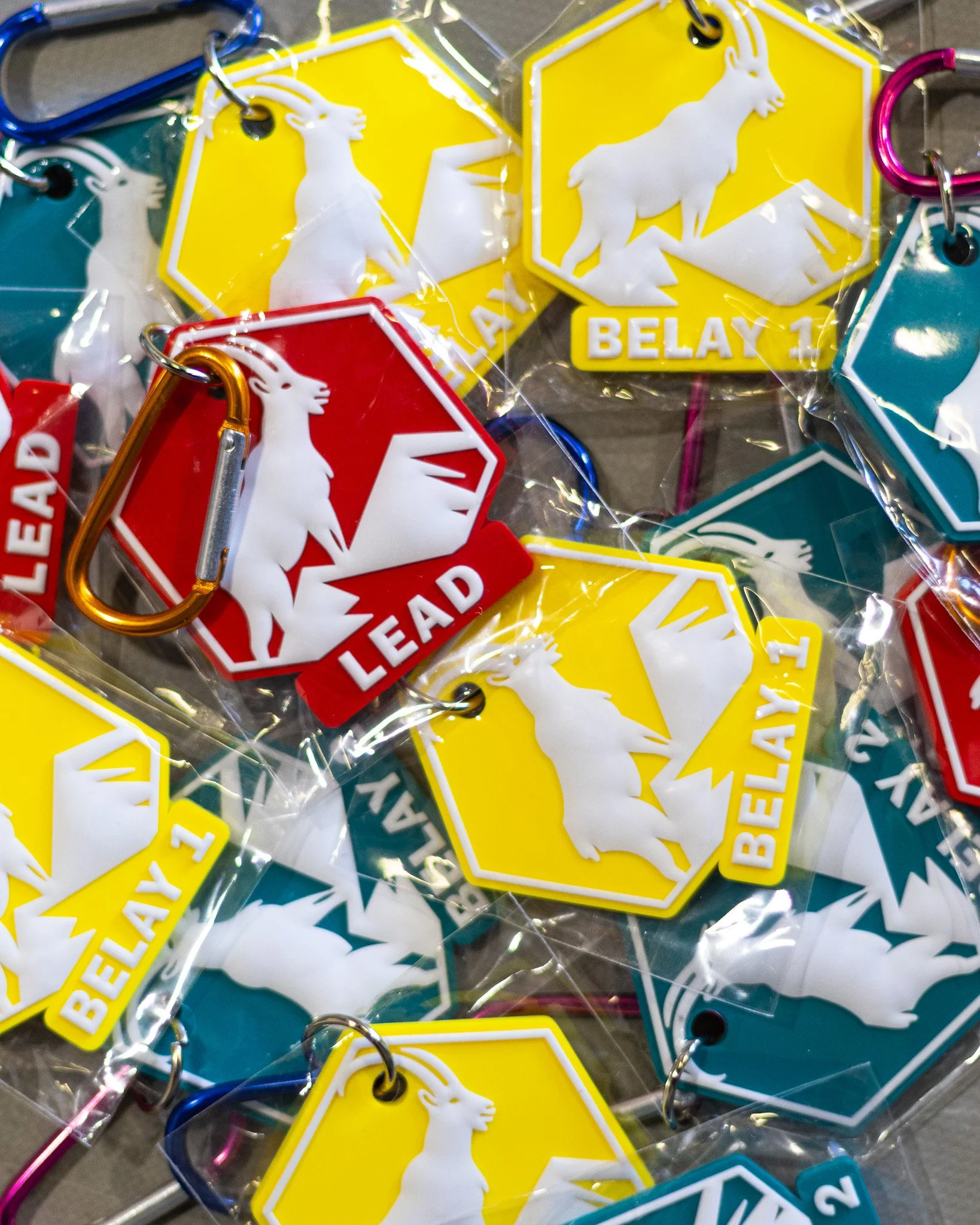 A pile of colorful keychains shaped like badges or tags with a white goat silhouette and the words 'LEAD' and 'BELAY' on them. Some keychains are attached to carabiners.