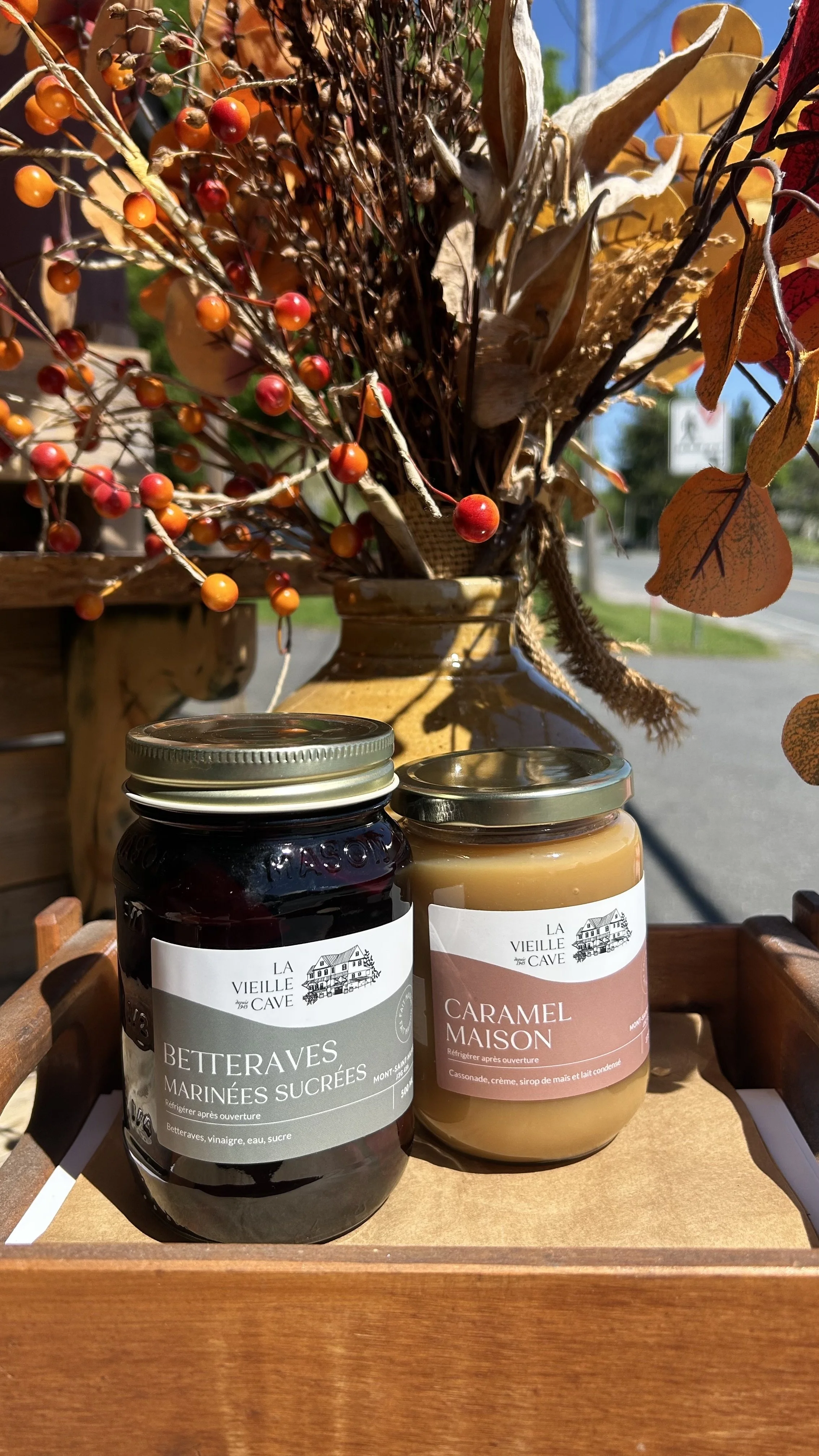 Deux bocaux de confiture, l'un de betteraves marinées sucrées et l'autre de caramel maison, placés sur une surface en bois. En arrière-plan, un bouquet de branches avec des baies rouges, des feuilles séchées et une décoration en céramique.