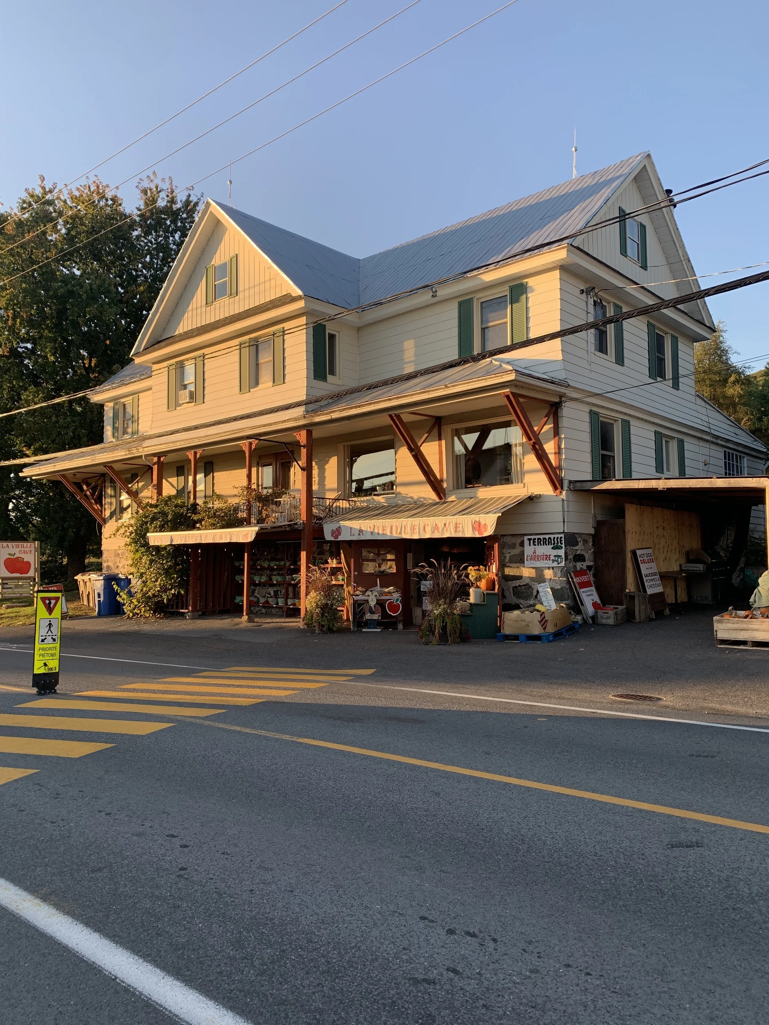Une maison à plusieurs étages avec une façade blanche et un toit en métal, située au bord d'une rue avec une marquage au sol jaune. Il y a un panneau de signalisation pour les piétons et divers panneaux et étalages en vente devant la maison.