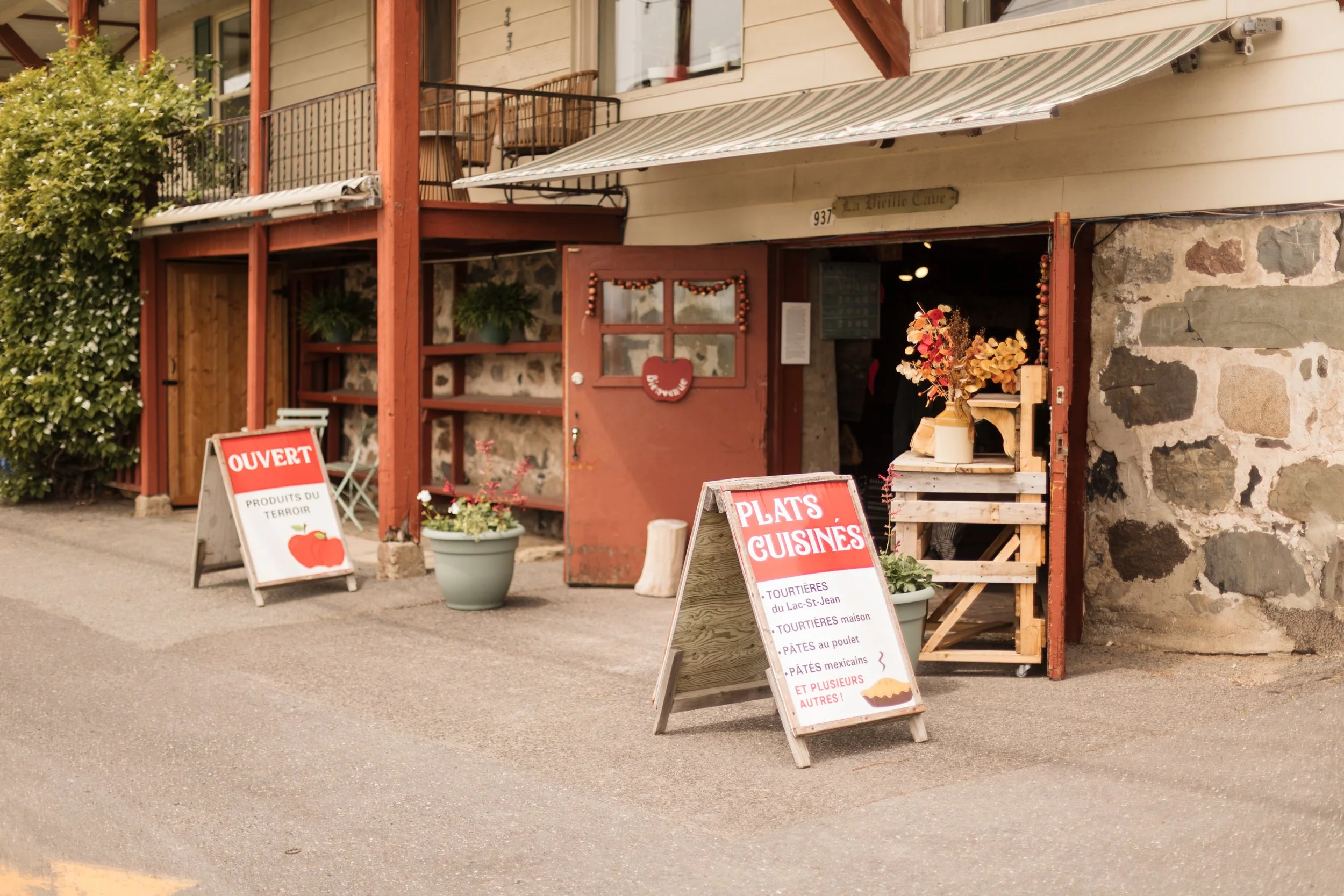 Façade d'une petite boutique en pierre avec devanture en bois rouge, proposant des plats cuisinés, avec des panneaux indiquant 'OUVERT' et des spécialités régionales en français.