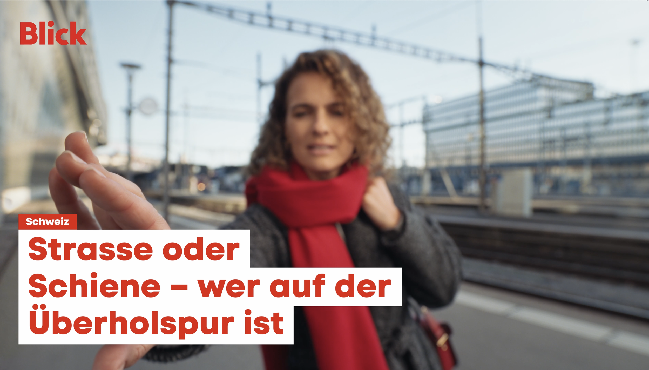 A woman with curly hair, wearing a gray coat and red scarf, holding out her hand as if to stop or signal, standing on a train platform with train tracks, overhead power lines, and a large building in the background.