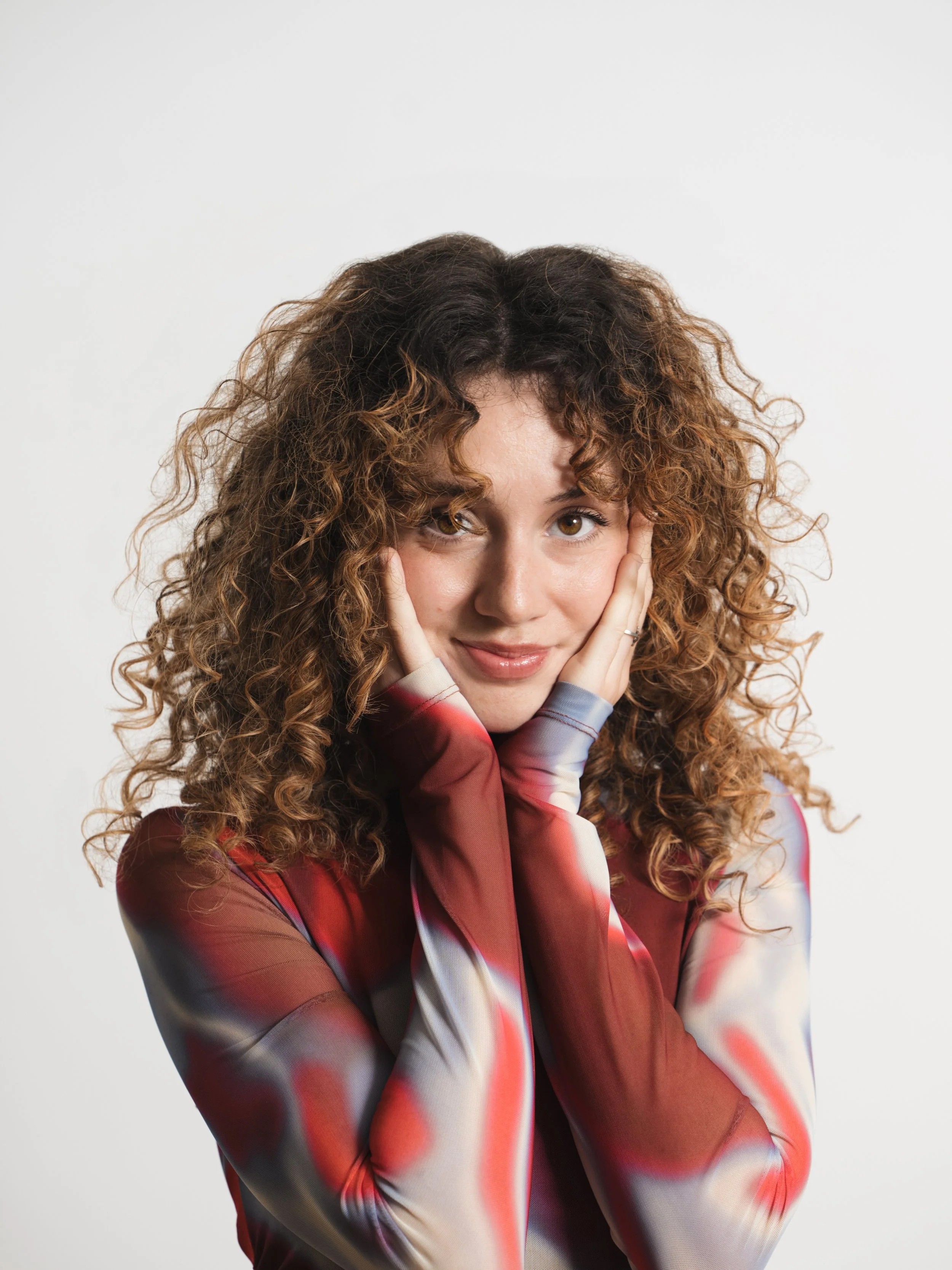 A woman with curly brown hair and light skin, smiling softly, holding her face with both hands, wearing a long-sleeve multicolored top with red, gray, and white patterns, against a plain white background.