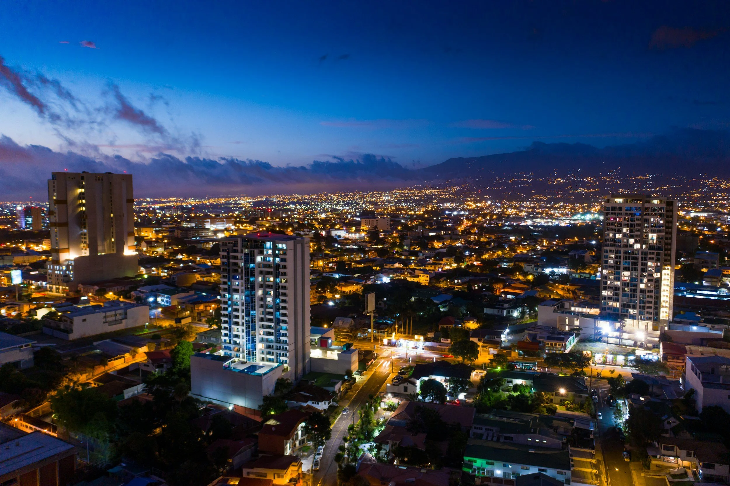 Nighttime cityscape with illuminated buildings and streets, a mountain range in the background, and a partly cloudy sky.