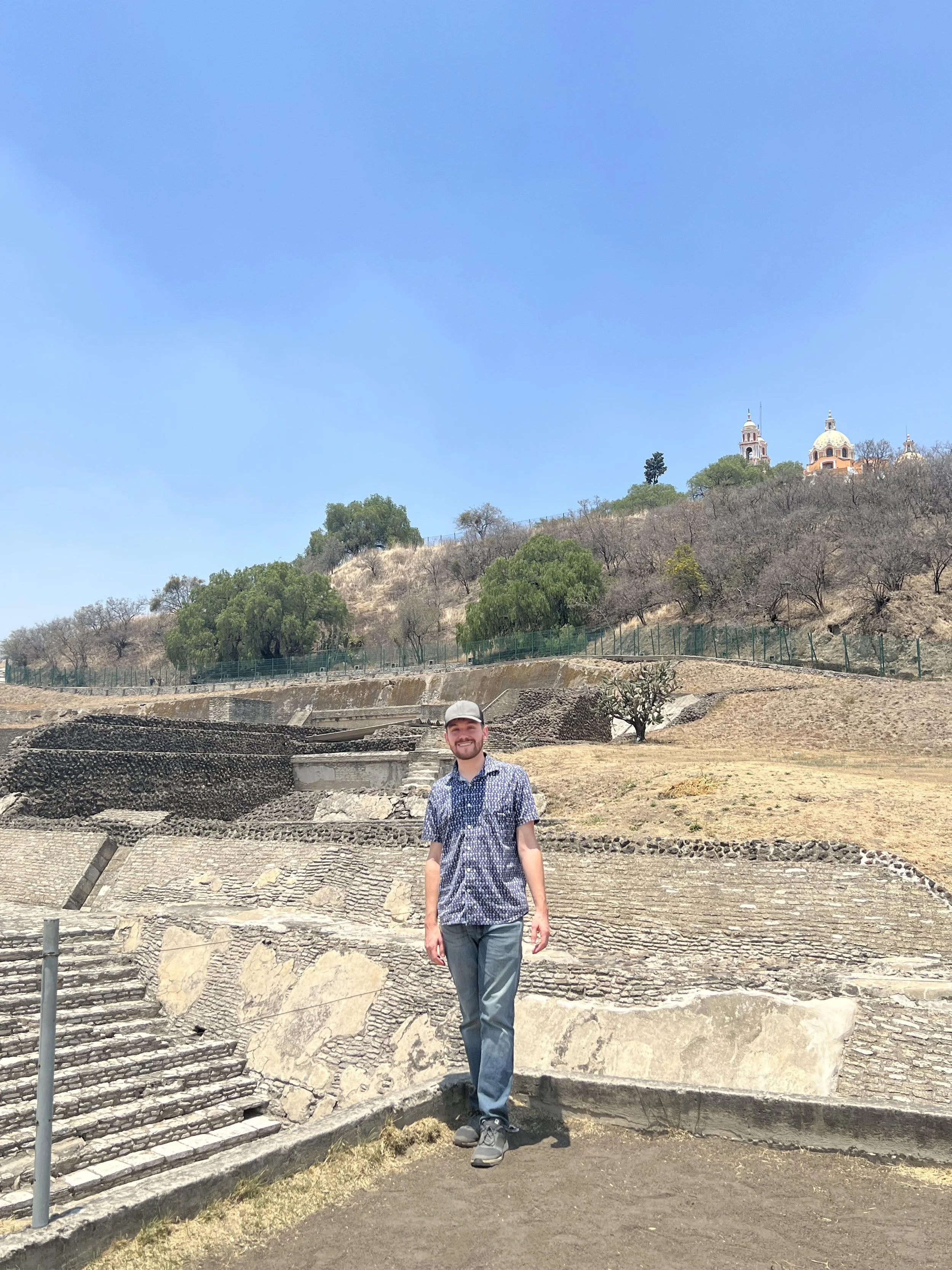 A man standing on ancient stone archaeological site with stairs, hills, trees, and buildings in the background on a sunny day.