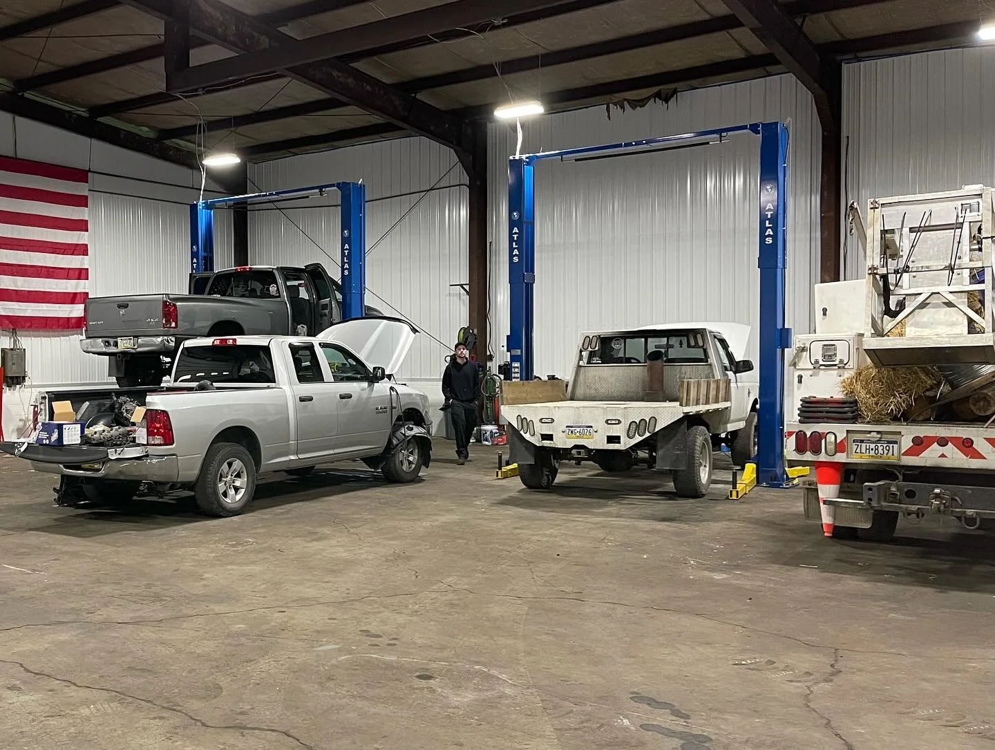 Inside an auto repair shop with two car lifts, one lift supports a curbside pickup truck, and the other lift supports an older flatbed truck. A man stands near the center, wearing a black jacket and cap, with an American flag hanging on the left side of the wall.