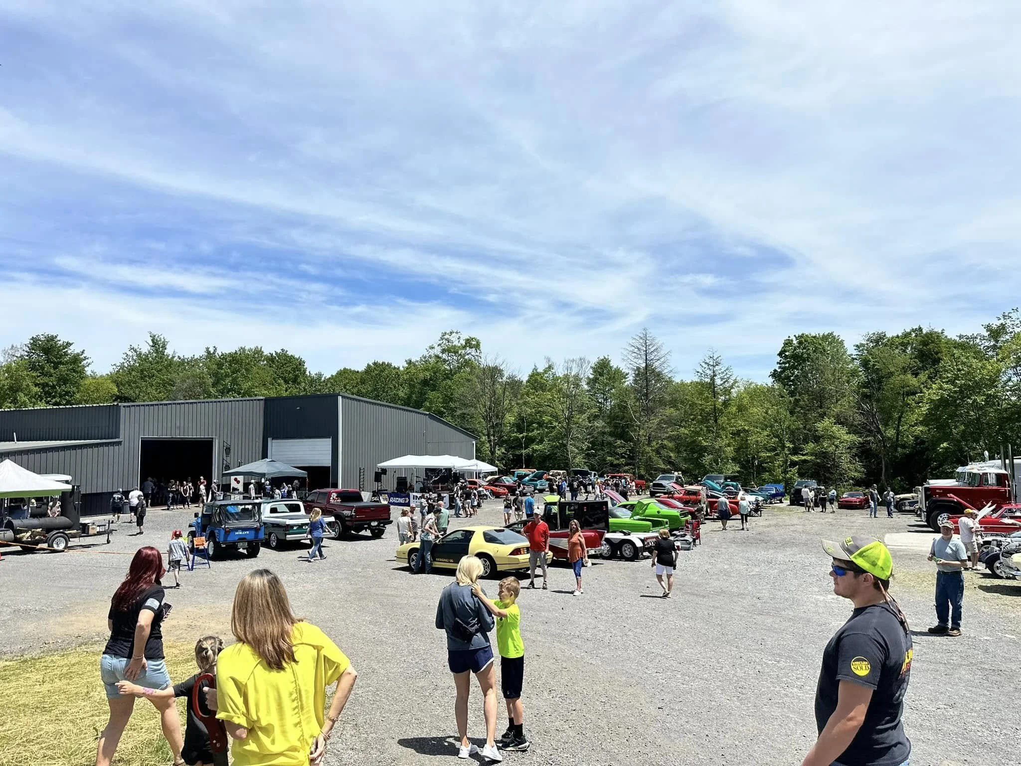 Outdoor car show with vintage and sports cars displayed in front of a large gray building, with people walking around, talking, and viewing cars on a sunny day with blue sky and trees in the background.