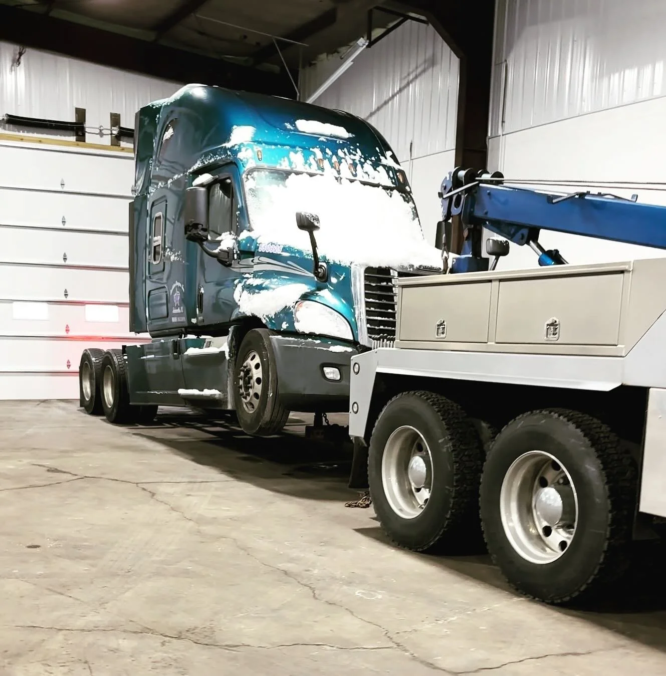 A large blue semi-truck with snow on the front is inside a warehouse, being serviced with a blue crane attached to a white service truck.