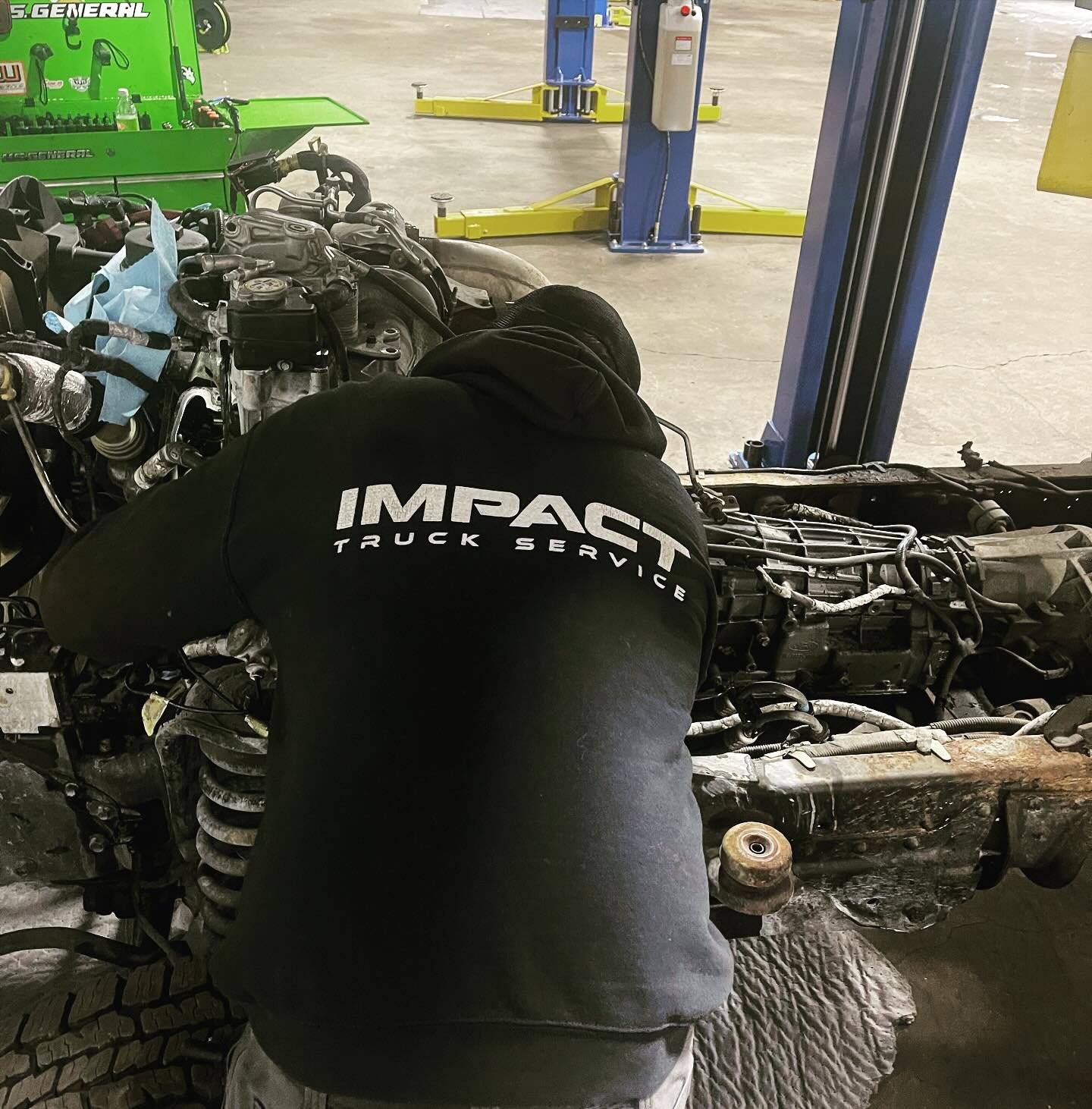 A mechanic working on a vehicle in a garage, wearing a black hoodie with "IMPAC Truck Service" printed on the back. The vehicle's engine and components are exposed, and other tools can be seen in the background.
