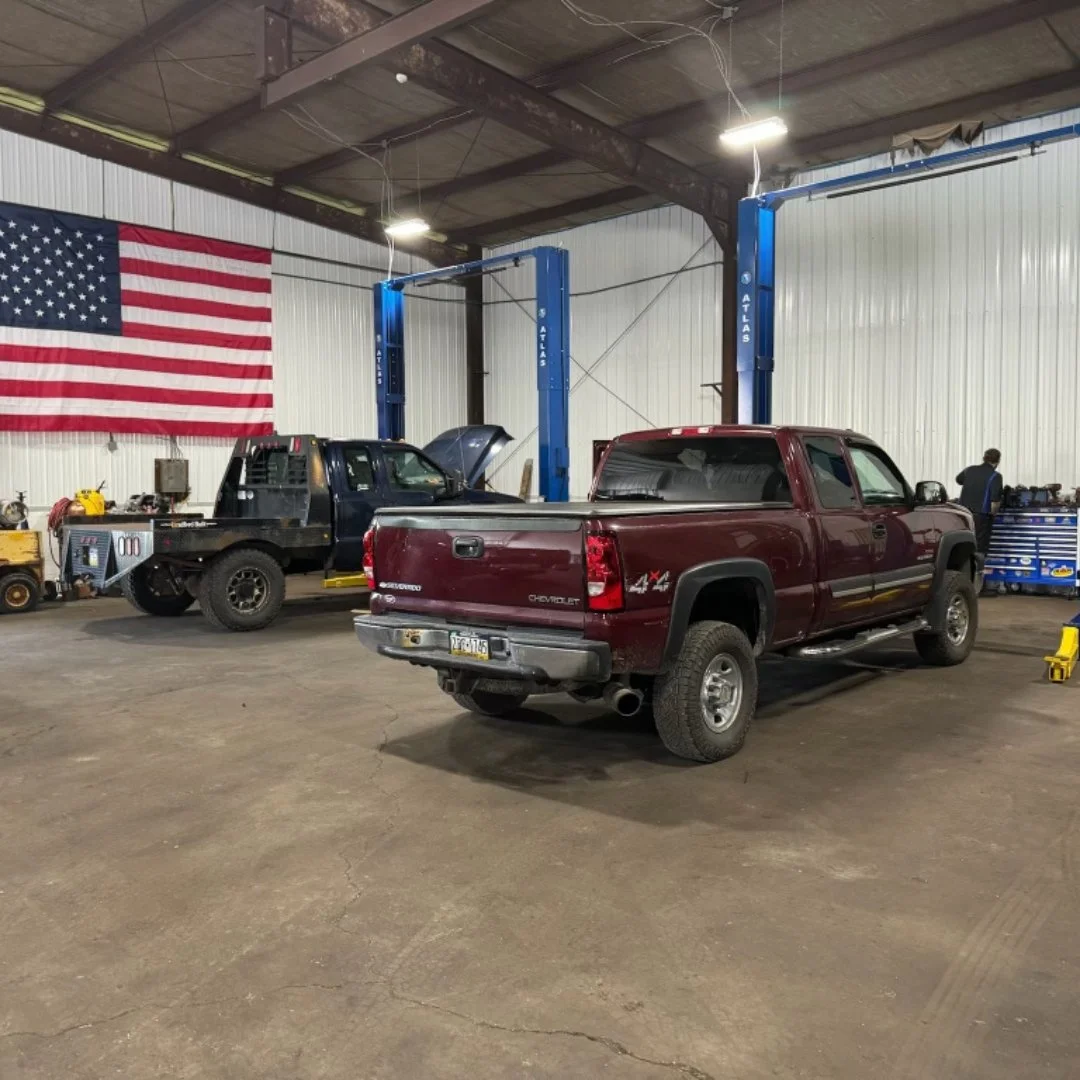 Inside a mechanic shop with a red Chevrolet pickup truck and a black vehicle with its hood open, a blue hydraulic lift, an American flag on the wall, and mechanic tools and equipment.