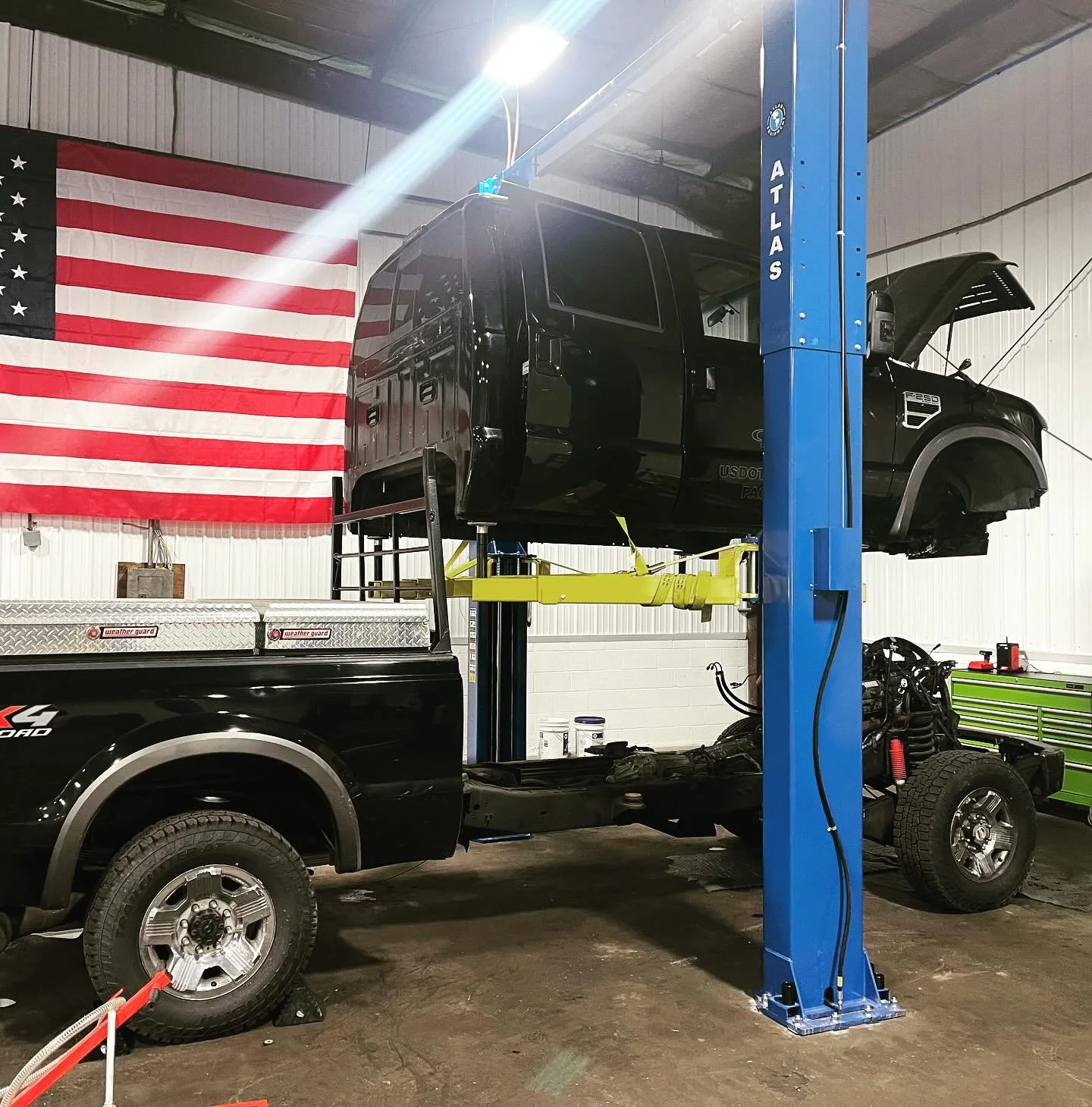 A black pickup truck with its cab lifted on a blue hydraulic lift inside an auto repair shop. The truck's bed is removed, exposing the chassis. An American flag hangs on the wall in the background.