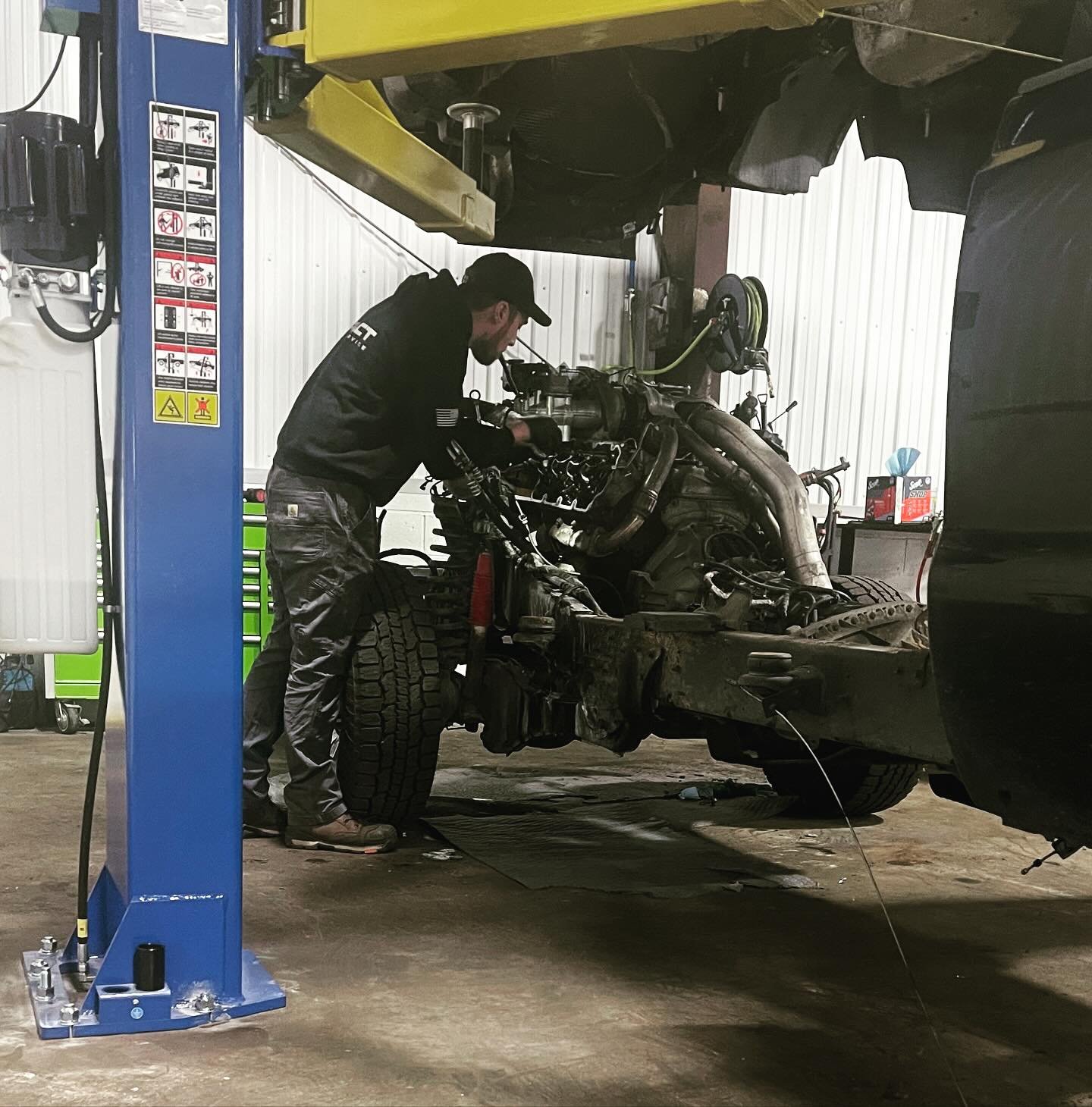 A mechanic working on the engine of a large vehicle, possibly a truck, in a garage or workshop. The vehicle is raised on a blue hydraulic lift, with the front of the vehicle exposed and the engine parts visible.