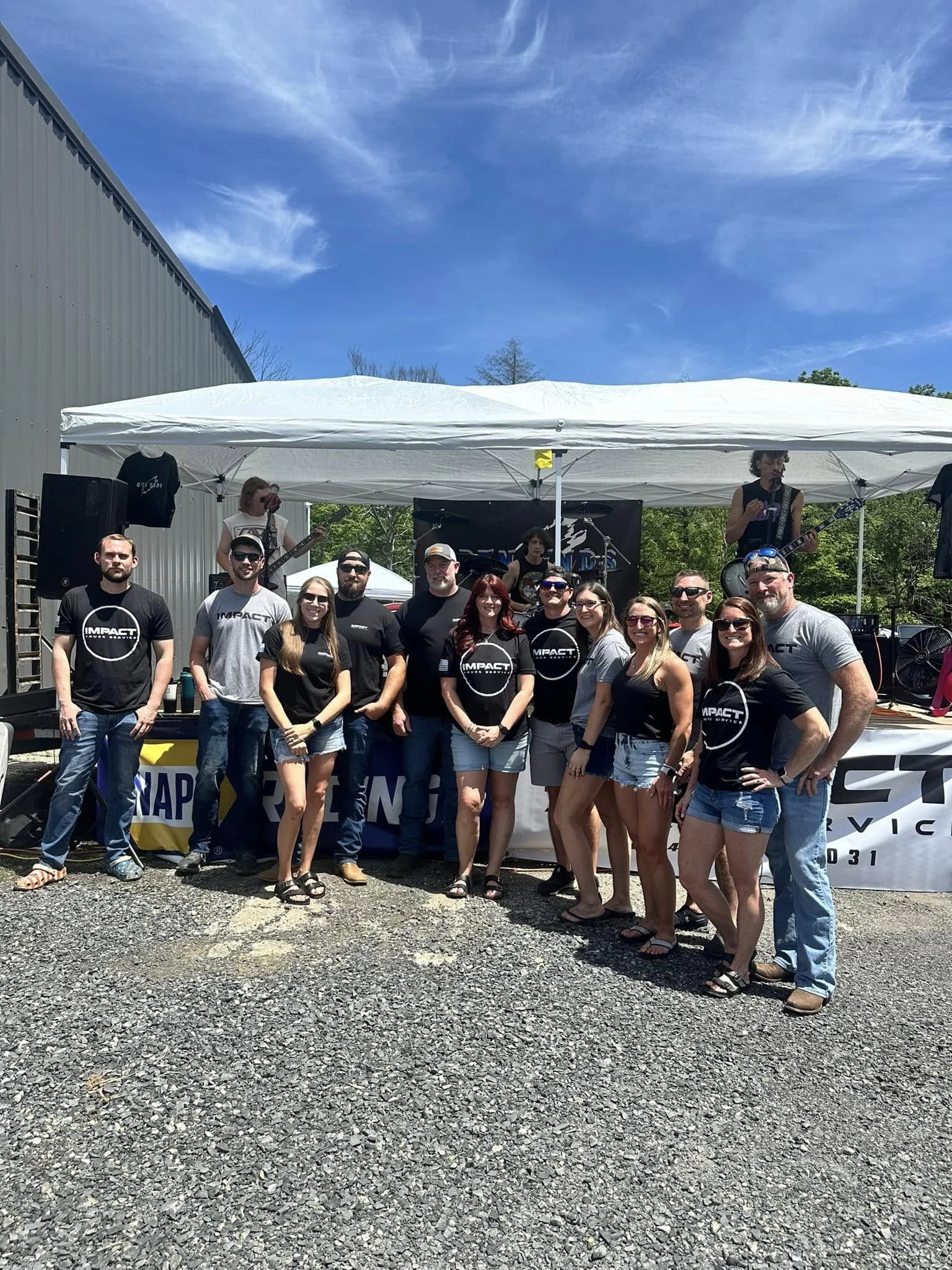 Group of people in front of a band performing on stage outdoors on a sunny day. The band members are playing instruments under a white canopy, with effects speakers and banners visible.