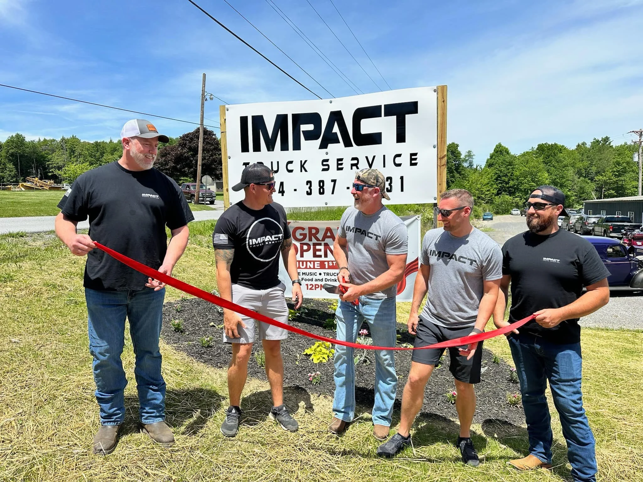 Five men standing outdoors in front of a sign for Impact Truck Service, participating in a ribbon-cutting ceremony, with one man holding scissors. All are smiling and wearing Impact shirts or hats, with a clear sky and greenery in the background.