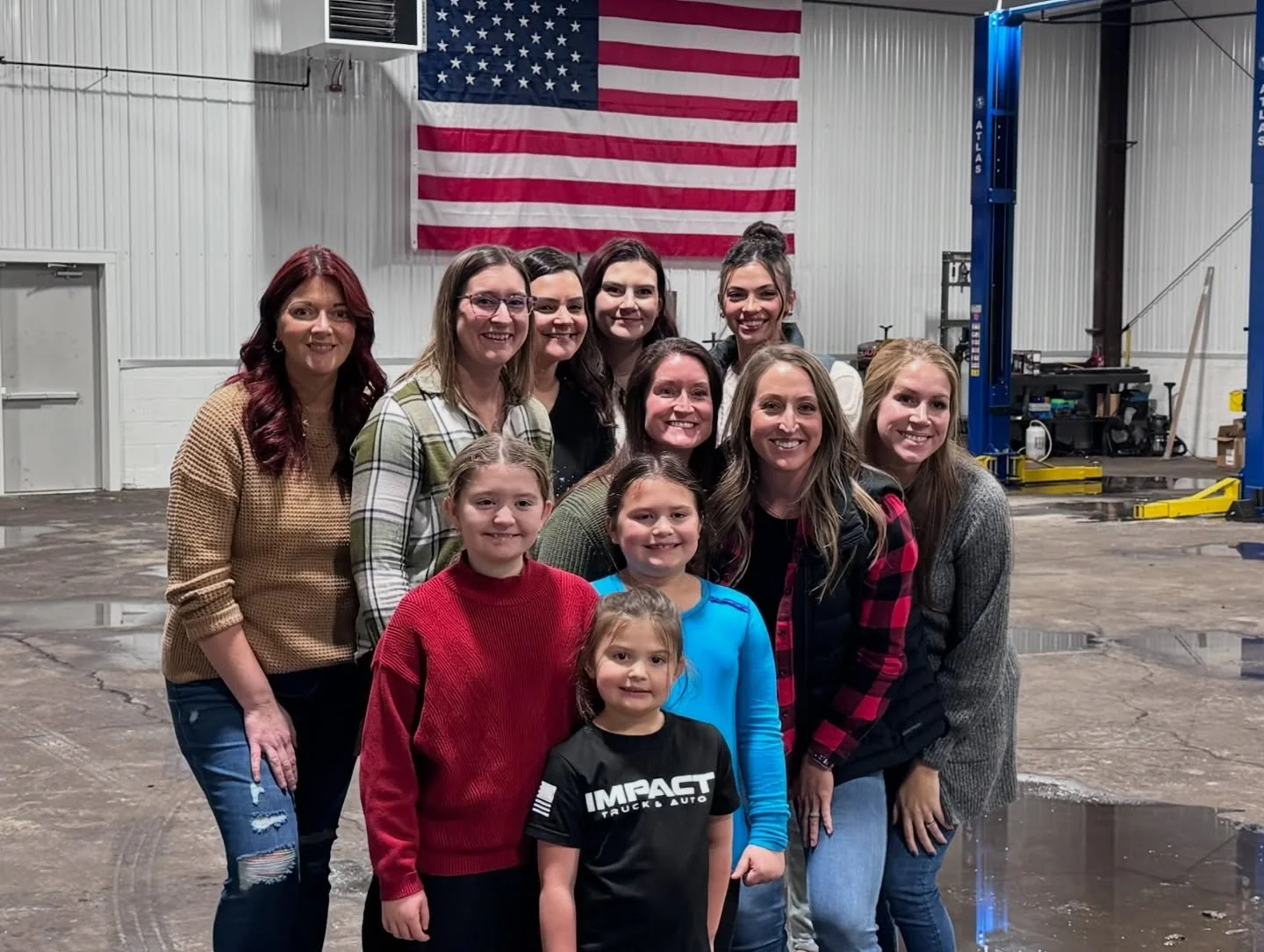 Group of ten women and girls posing inside a large industrial space with an American flag hanging on the wall in the background.