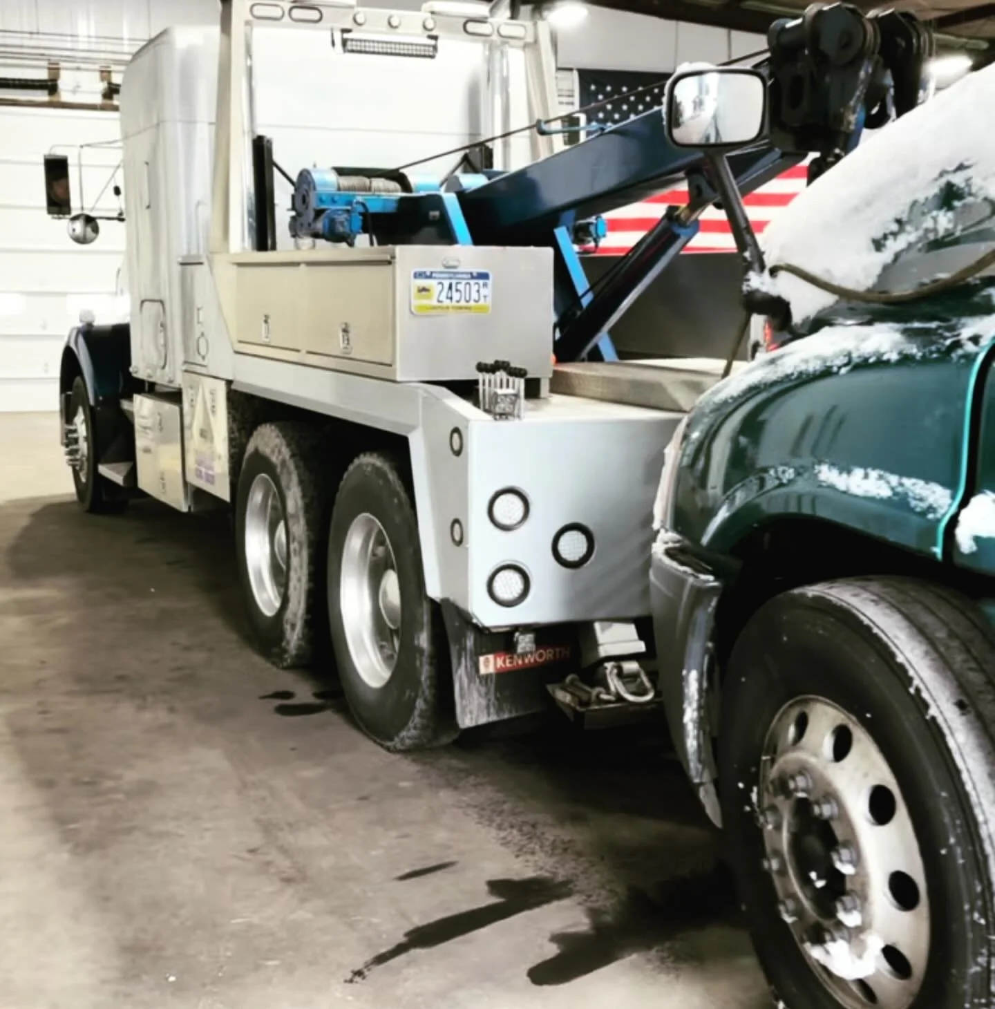 A tow truck parked in a garage, with a partially snow-covered green vehicle attached. The tow truck has multiple axles and a lift mechanism on top, with some snow on its components.